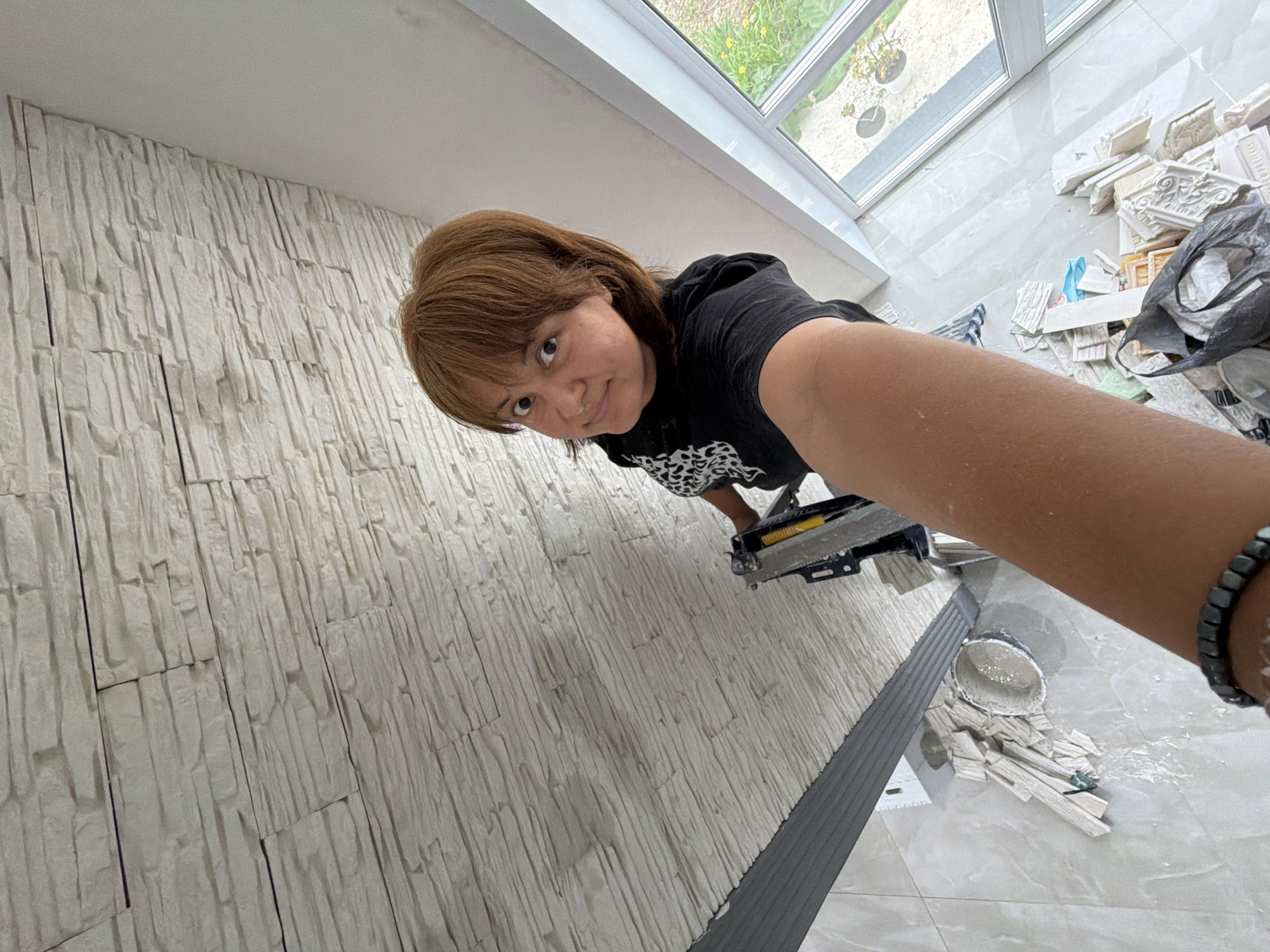 A woman with shoulder-length brown hair is taking a selfie from a high angle. She is wearing a black hoodie with a white graphic design on it. She is standing on a ladder, as a power tool with a yellow handle is visible next to her arm, and she is on a ladder or scaffolding. The background shows a wall covered in textured, light-colored stone tiles, extending upwards and to the left. To the right, a large window reveals a glimpse of the outdoors with greenery and a potted plant. Below, on the floor, there are scattered pieces of the same stone tiles, a white bucket with some liquid inside, and a black plastic bag filled with what appears to be building materials. The floor is tiled with large, light-colored tiles. The scene suggests renovation or construction work is in progress.
