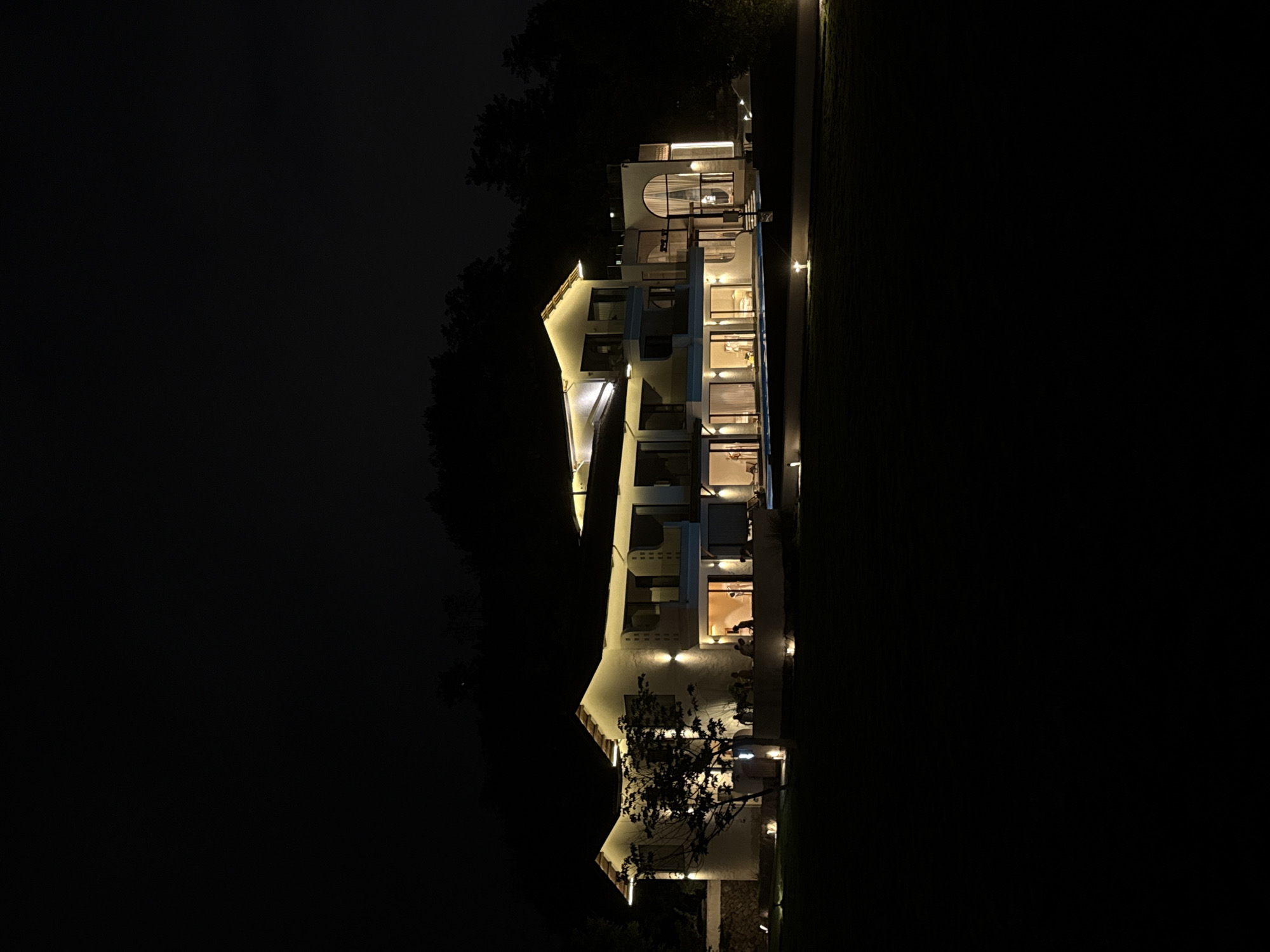 A multi-story white building is illuminated at night, with lights on both the exterior and visible within the windows. The building is situated between dark trees and a dark grassy area. The location is likely in Jinhua, China, given the request. No people are directly visible, though there are indications of occupancy through interior lights. The environment is dark, with artificial light sources providing illumination.