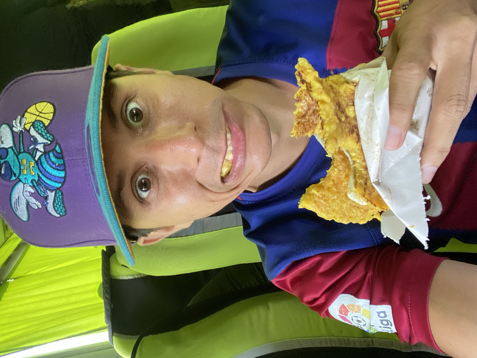 A man with light skin and short dark hair wears a purple baseball cap with a cartoon logo and a Barcelona soccer jersey. He is holding a yellow food item wrapped in white paper in his right hand and appears to be eating it. The background appears to be a bus seat with a green cover. No further information about La Morita, Venezuela is discernible from the image.