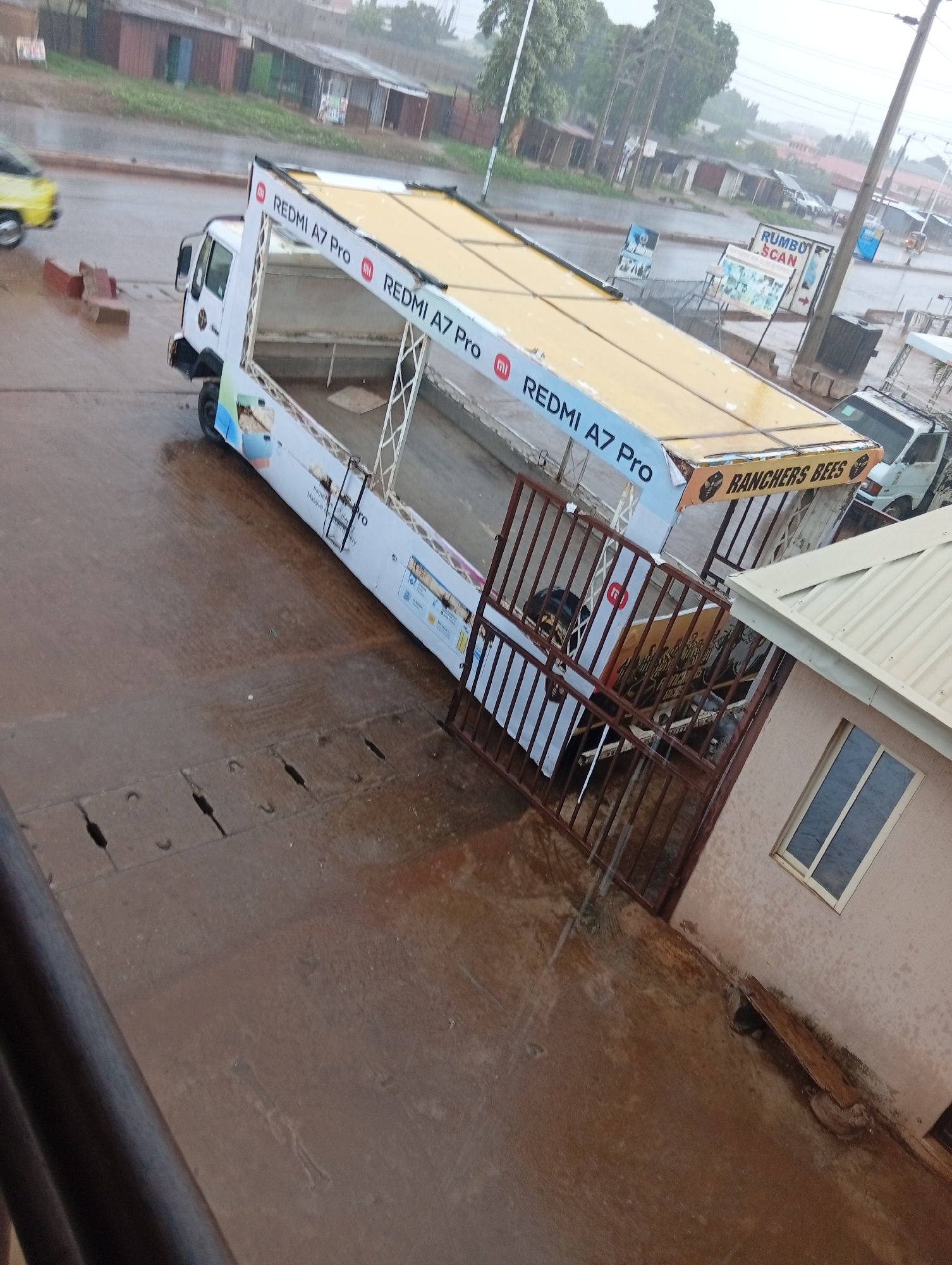 The image captures a wet scene in what is identified as Sabon Tasha, Nigeria. A white truck dominates the frame, featuring the branding "REDMI A7 Pro" on its canopy and sides. Additional branding, “RANCHERS BEES,” appears on the rear of the canopy. The truck is parked beside a low, single-story building with a window. A metal gate separates the truck from the building. The street is wet, suggesting recent rainfall. Other vehicles are visible in the background, along with signage for "RUMBOT SCAN." The scene gives the impression of an urban or suburban area in the midst of a downpour.
