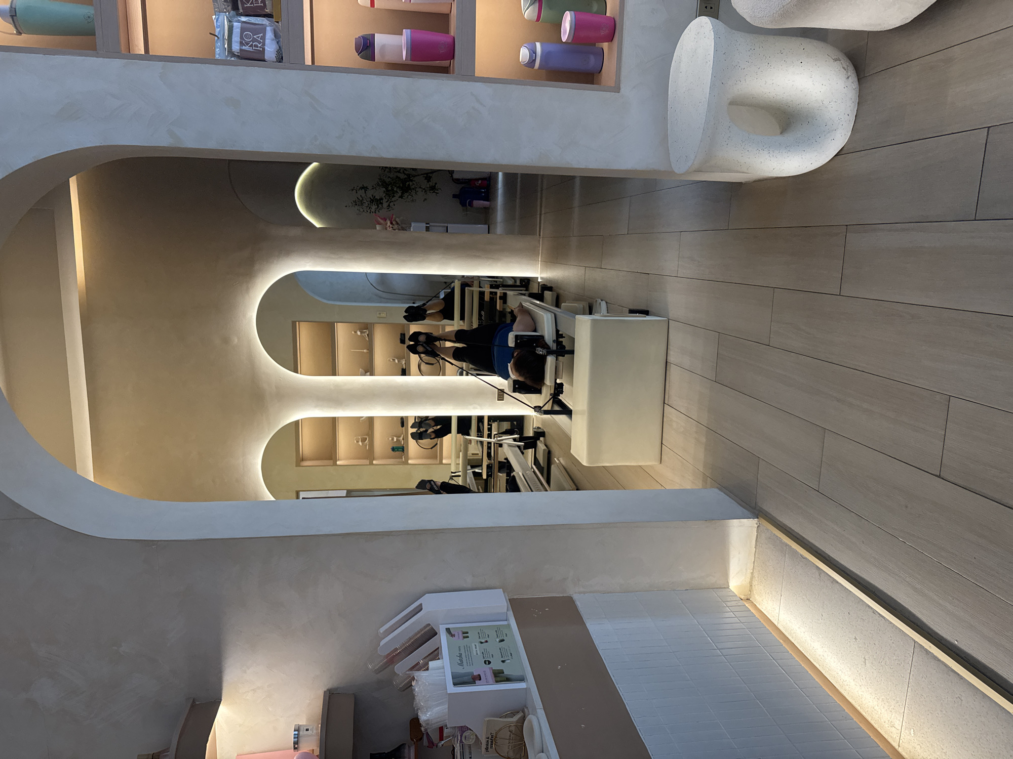 The image depicts the interior of a Pilates studio. Two individuals are engaged in Pilates exercises on reformer machines. The studio has a modern design with curved archways and recessed lighting. Various workout tools and bottles of merchandise are displayed on shelves. The flooring is tiled. The location may be in Davao City, Philippines.