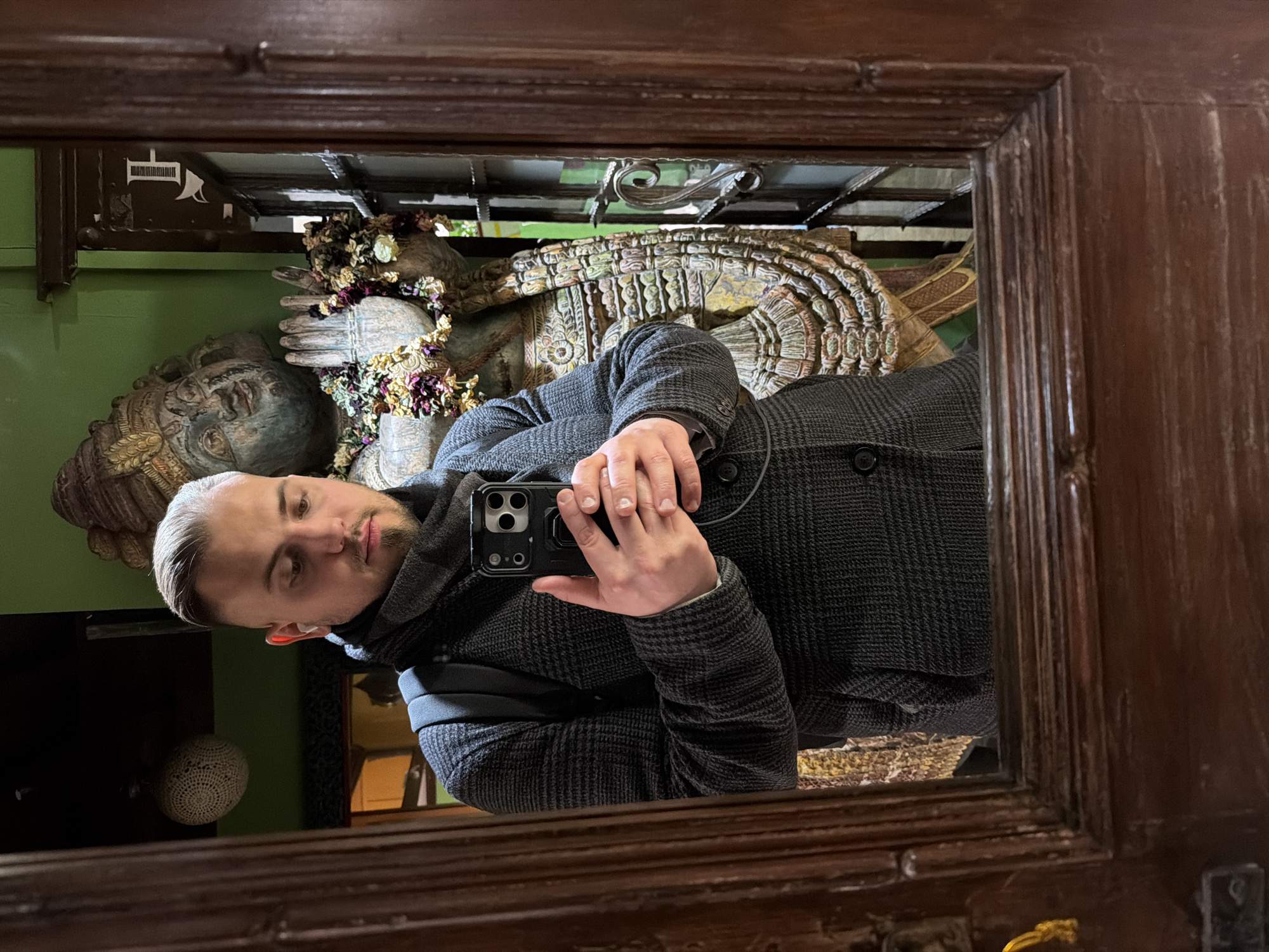 A man wearing a gray patterned coat is taking a selfie in a mirror. Behind him, reflected in the mirror, are religious statues with floral decorations, suggesting a temple or place of worship. The setting appears to be in Moscow, Russia, due to the architectural style and religious artifacts present.