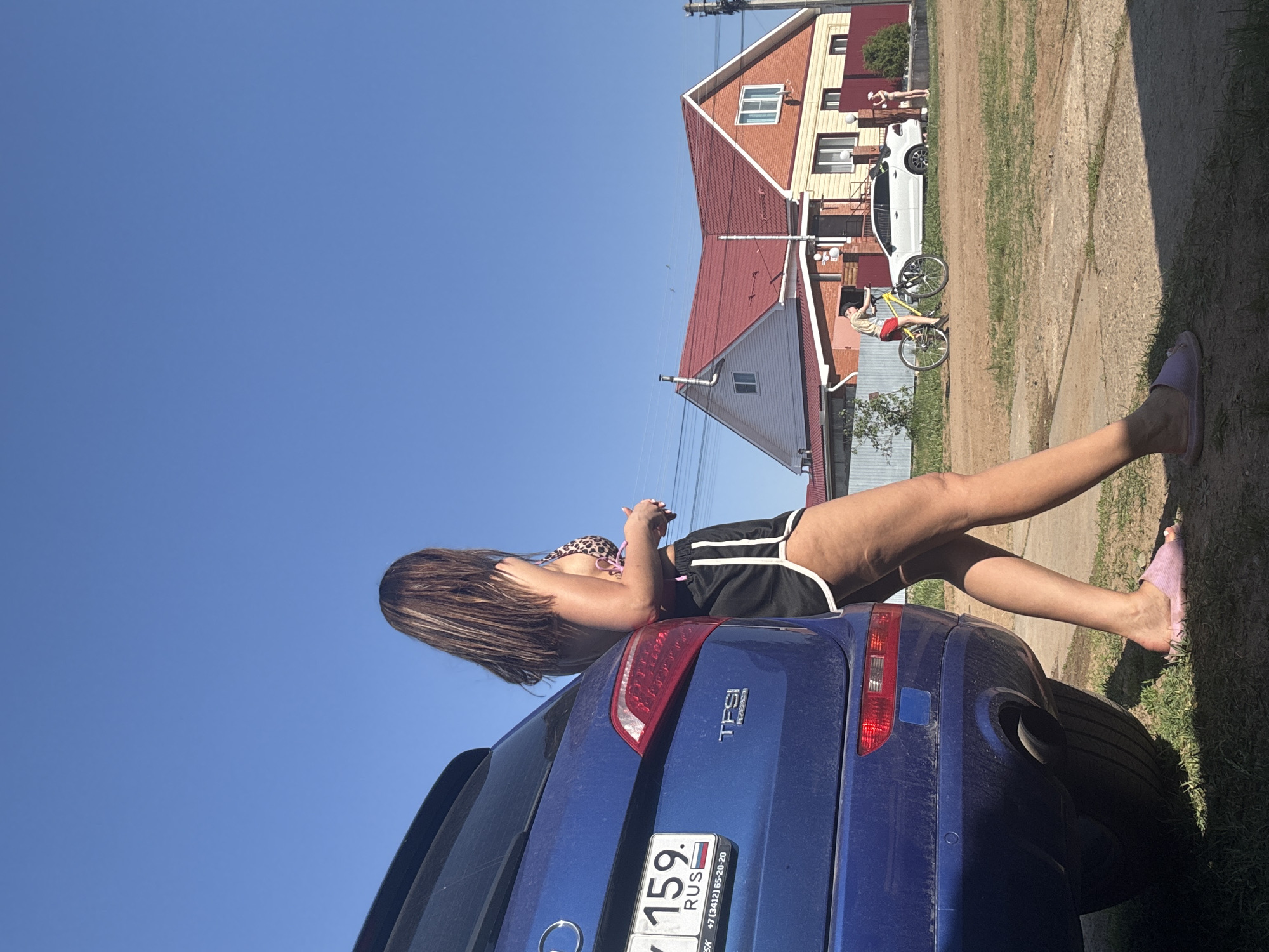 A medium shot, angled upward, depicts a young woman reclining on the back of a blue SUV, possibly an Audi, judging by the "TFSI" badging. She is wearing a bikini top with an animal print and black shorts with white trim. Her dark, streaked hair is partially obscuring her face as she appears to be looking at or interacting with something in her hands. A Russian license plate is visible on the vehicle, beginning with "7," and is mounted in the city of Dubovaya, Russia.
In the background, a red-roofed house with brick siding is seen, along with a white car and two individuals; one walking and another riding a bicycle. The scene suggests a suburban or rural setting with a clear blue sky above. The ground is a mix of grass and dirt.