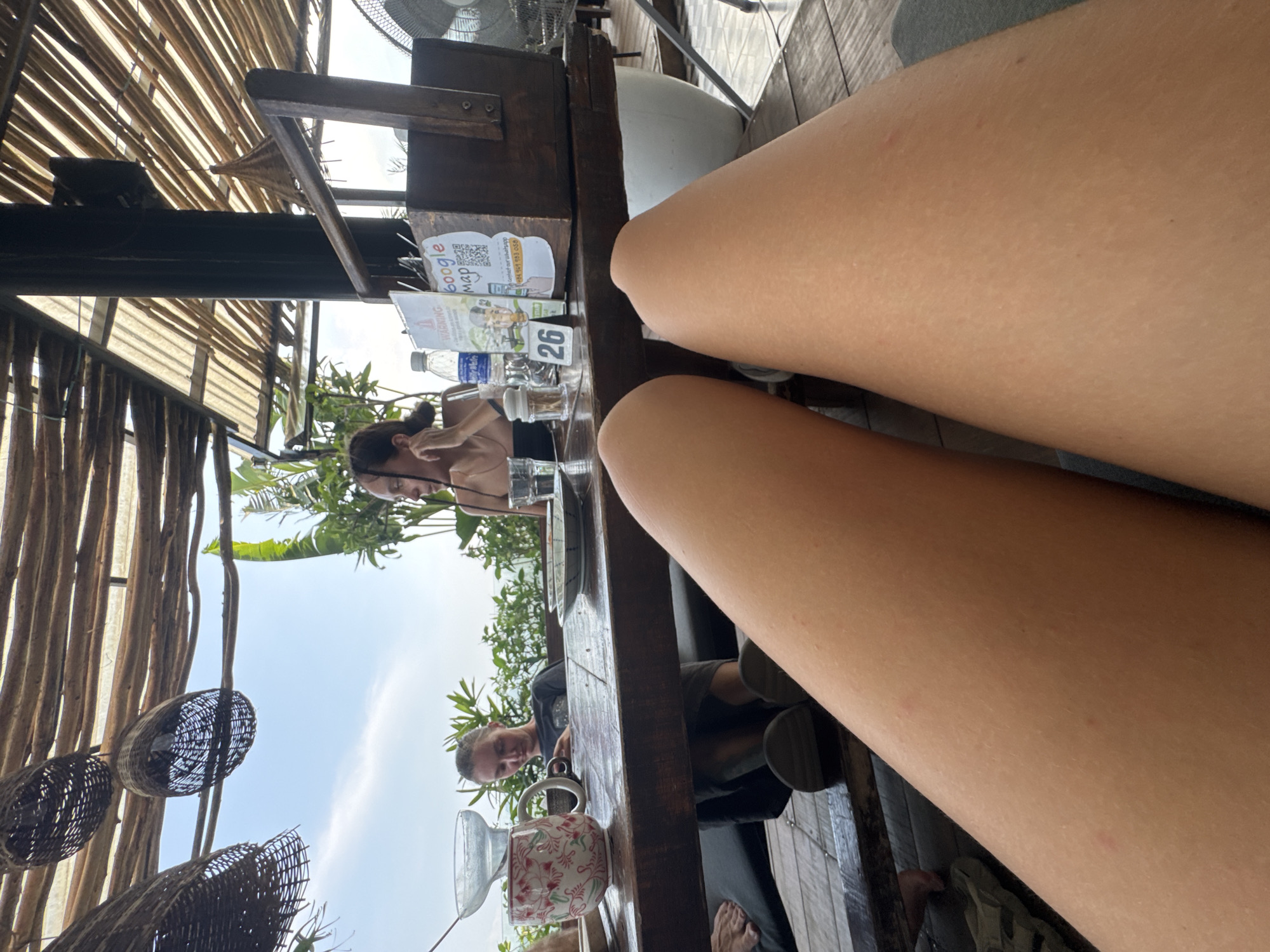 The image captures an outdoor dining scene from a first-person perspective, with the photographer's bare legs extending into the immediate foreground, resting on or near a rustic wooden table.
On the table, various items are visible: two clear plastic water bottles, multiple drinking glasses, a small white mug adorned with a red floral pattern, and a salt and pepper shaker. Information cards are placed on the table, one displaying "google map" with a QR code and a phone number beginning with "+84", and another with the word "WARNING". A small stand clearly shows the table number "26".
Two other individuals are seated at the table. To the left, a woman with dark hair tied back leans forward, appearing engaged with something on the table. Further down, another person is partially visible, also seated.
The setting is an open-air establishment, likely a cafe or restaurant in Hà Giang 1 Ward, Vietnam. It features a slatted bamboo or wooden roof structure providing shade, from which woven basket decorations are suspended. The background shows a clear, bright sky and lush green foliage, indicative of a warm, tropical climate. A ceiling fan is partially visible overhead. The bright daylight suggests the time is likely daytime, either morning or afternoon. The skin on the visible legs appears fair with a few small red marks.