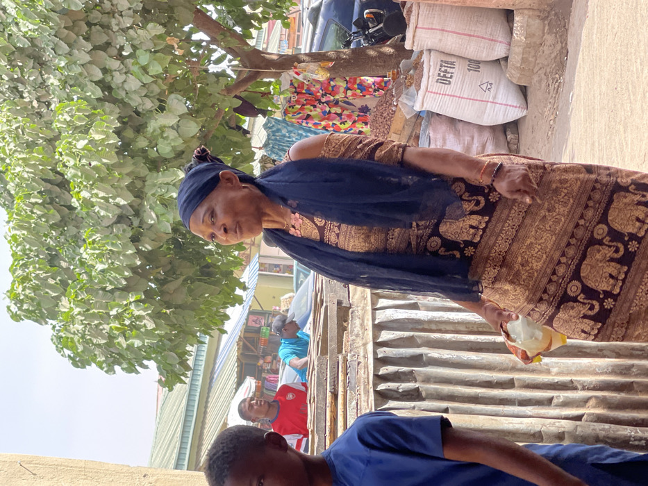 An outdoor market scene is depicted in Samaru, Nigeria, during daylight hours. In the mid-ground, an adult female with dark skin is positioned slightly to the right of the frame's center, looking upward and to the right with her mouth slightly open. She wears a black headwrap, a black sheer scarf, and a long brown and gold dress patterned with elephant motifs. A pink beaded necklace is visible, along with an orange/red beaded bracelet on her left wrist. She holds a yellow and white plastic item in her left hand at hip level.
To her immediate left, in the foreground, the left side of a young male's head and blue t-shirt are partially visible. Behind the main female subject, an adult male in a red t-shirt with a white logo is visible in the mid-background, looking right. Further left and behind him, another adult male in a bright blue t-shirt and a dark cap leans over a corrugated metal and wooden structure, possibly a market stall.
To the right of the main female subject, a large tree with green foliage stands. Hanging from a line near the tree are various patterned clothing items. At the base of the tree, multiple large white sacks, two marked "DEFTA 100", contain what appears to be agricultural produce. A motorcycle is partially visible behind these sacks. The background features several market stalls with corrugated roofs, other individuals, and parked vehicles, indicating a lively commercial area. The sky above is clear, suggesting bright weather conditions.