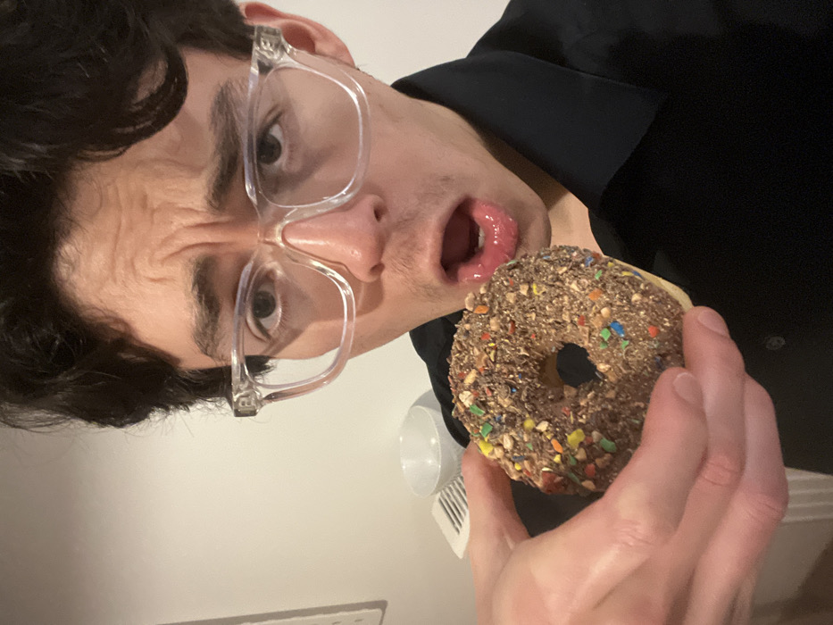 A man with dark hair and clear glasses holds a chocolate donut covered in sprinkles. His mouth is open, tongue slightly visible, appearing ready to eat the donut. He wears a black shirt. The background shows a white wall and a partial view of a white object. The picture is rotated.