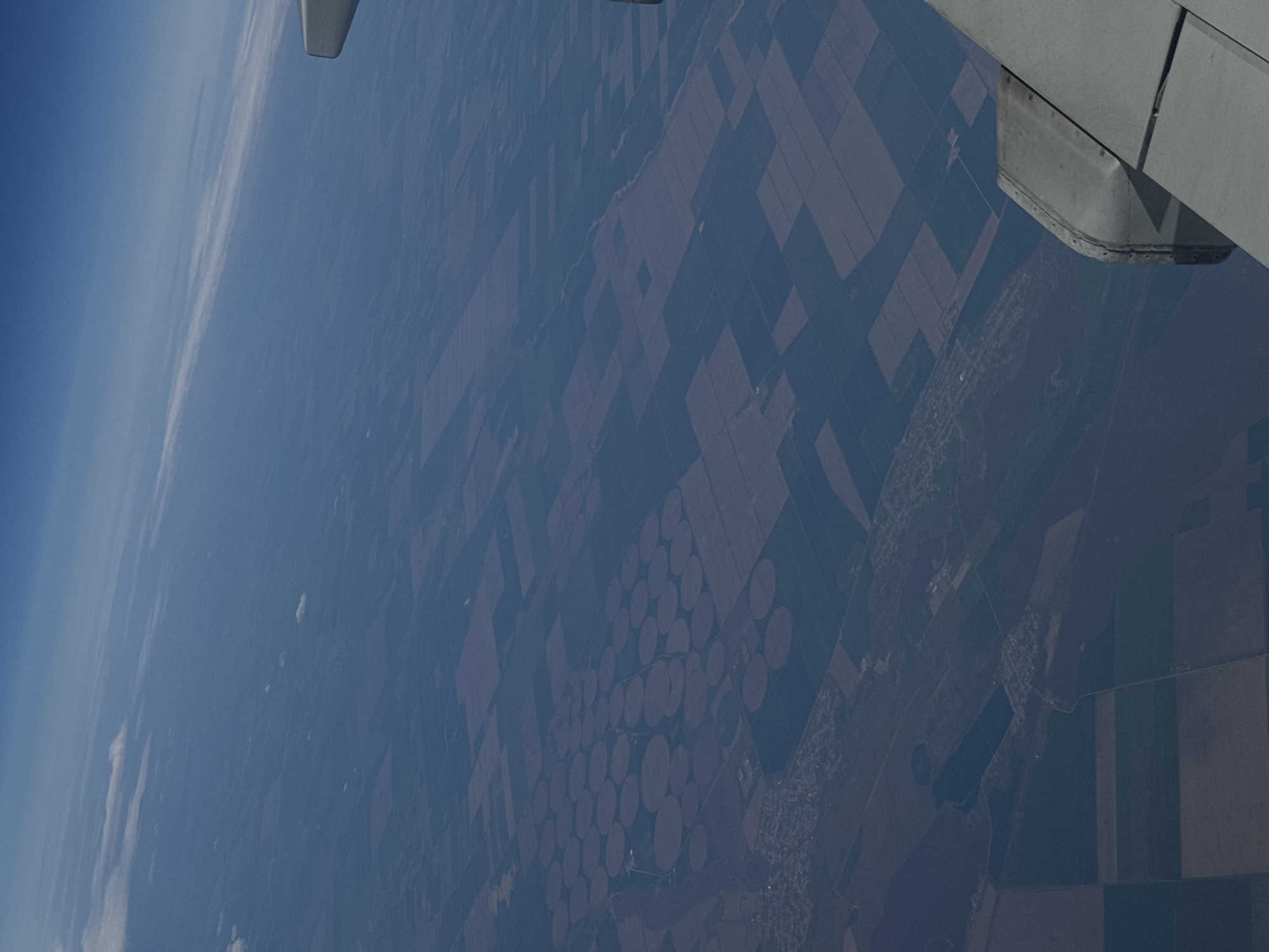An aerial view from an airplane over Забегалово, Russia, reveals a landscape dominated by agriculture. The earth below is patterned with numerous rectangular fields and circular shapes, indicative of center-pivot irrigation systems. An aircraft wing is visible in the upper right corner of the frame. The atmosphere is somewhat hazy. The scene contains no people or visible actions.