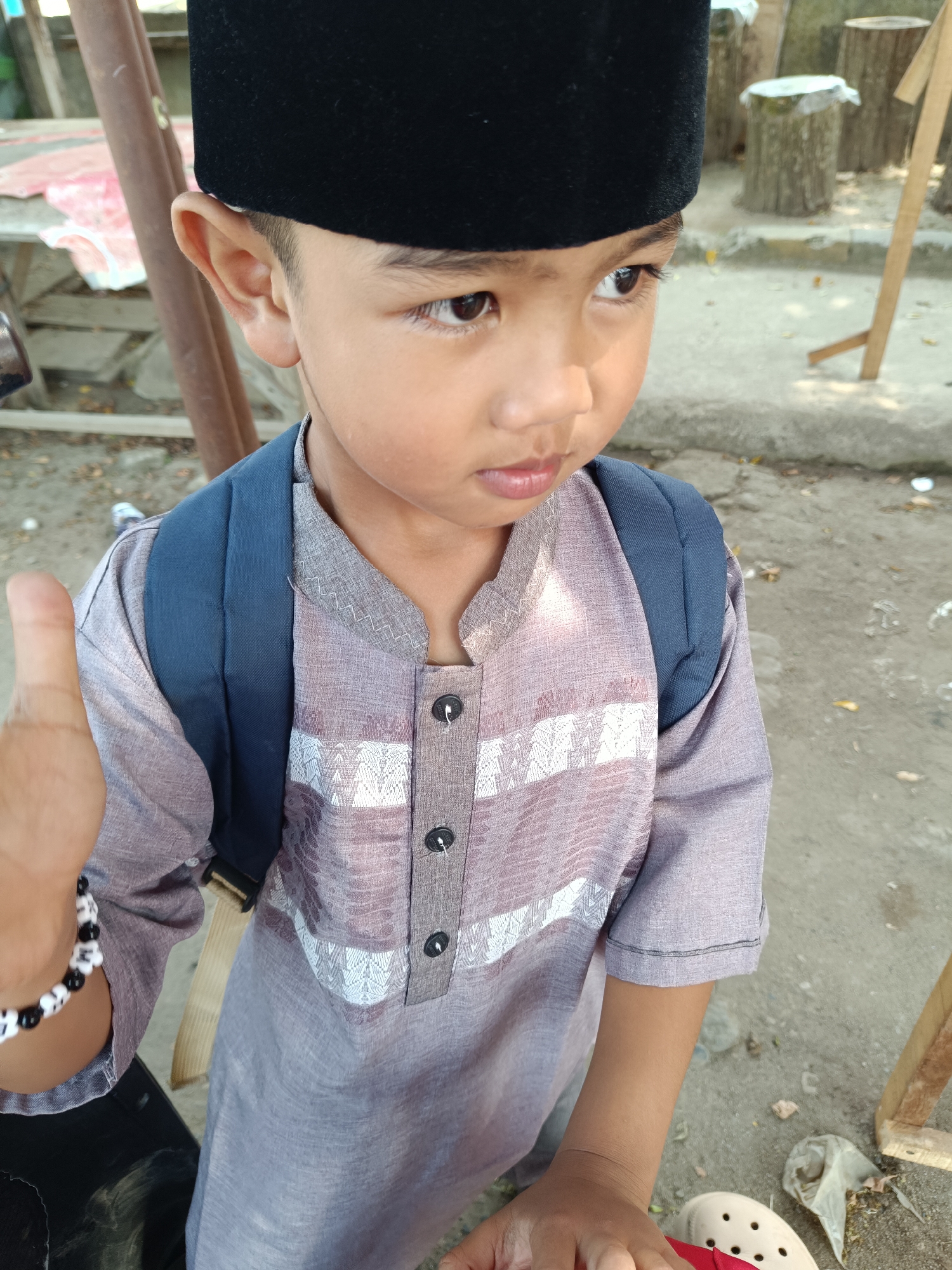A young boy is wearing a black peci and a light purple, long-sleeved shirt with white patterned accents and black buttons. He has a dark blue backpack on. His left arm is raised with his thumb up, and he is wearing a black and white beaded bracelet. He is looking to the right with his mouth slightly open. The background appears to be an outdoor area with concrete ground, some debris, and wooden structures. This scene is located in Way Timah, Indonesia.
