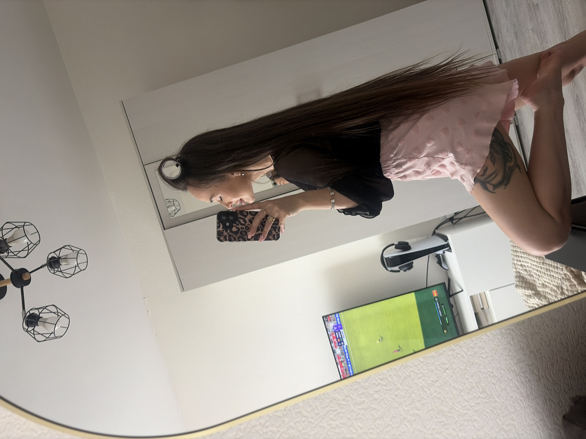 The scene is a full-body mirror selfie taken by a woman in a private residence, reportedly located in Saint Petersburg, Russia. The woman, who has long, straight, dark brown hair, is reflected in a large, oval-shaped mirror with a gold-colored frame, and is in a kneeling or crouched position on a light-colored floor, facing left in profile. She wears a sheer black long-sleeved top and light pink shorts with subtle heart patterns. A circular hair accessory is visible in her hair near the crown of her head. Her right hand, adorned with multiple rings, holds a smartphone with a leopard print case, directed towards the mirror to capture the image. A silver bracelet with a bear charm is on her left wrist. A prominent tattoo of a face or portrait is visible on her left upper thigh.
The room's reflection reveals light-colored walls, a modern black ceiling light fixture with four geometric caged bulbs, and a large white wardrobe. To the left of the wardrobe, a flat-screen television is displaying a football (soccer) video game in progress. Adjacent to the TV, a white PlayStation 5 console, a white gaming controller, and a matching white and black headset are arranged on a white cabinet. A textured, light-colored blanket or throw is visible in the lower left reflection, possibly indicating a bed or sofa. The wall behind the mirror has a subtle textured pattern.