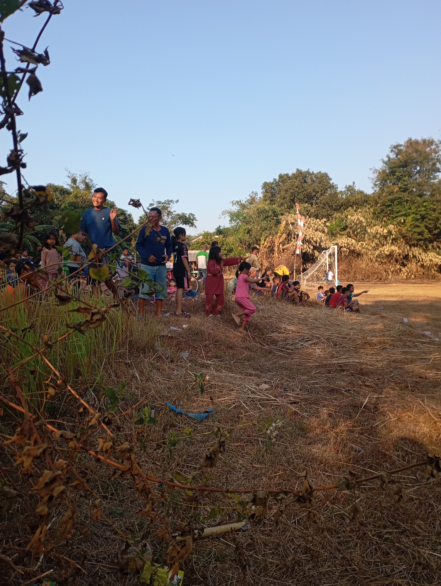An outdoor daytime scene depicts a group of people, primarily children, gathered in an open, dry, grassy field. In the foreground, dry, brown grass and branches of a dead plant are prominent. In the middle ground, a collection of people are seated and standing. Several children are playing or watching an activity. Some children are wearing colorful clothing, while others are in casual attire. A soccer goal with a white net is visible in the background, suggesting a sporting event or recreational area. Trees and foliage form a backdrop to the scene. The sky is clear and blue, indicating fair weather. The presence of a soccer goal and the informal gathering of people suggest this location is likely a public space or park within Kecamatan Haurgeulis, Indonesia, used for community events and recreation.