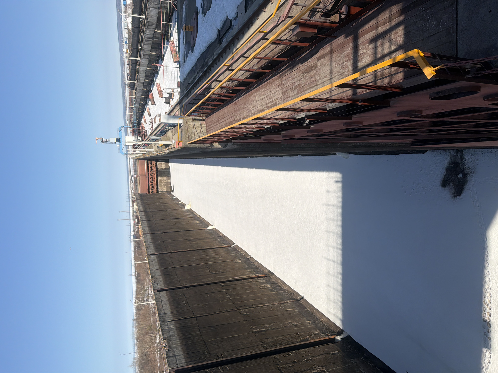 The image shows an industrial landscape in Chaikovsky, Russia, possibly depicting a frozen body of water, likely the Kama River, adjacent to industrial structures. The camera is positioned at an elevated angle, providing a view of the frozen water, a dark, wooden-slatted structure, and metallic industrial infrastructure with yellow railings. Snow covers the ground, and there is a clear, blue sky in the background. Details suggest an outdoor environment with industrial buildings and facilities along the river. The focus is on the frozen landscape and the surrounding industrial features. No people are visible.