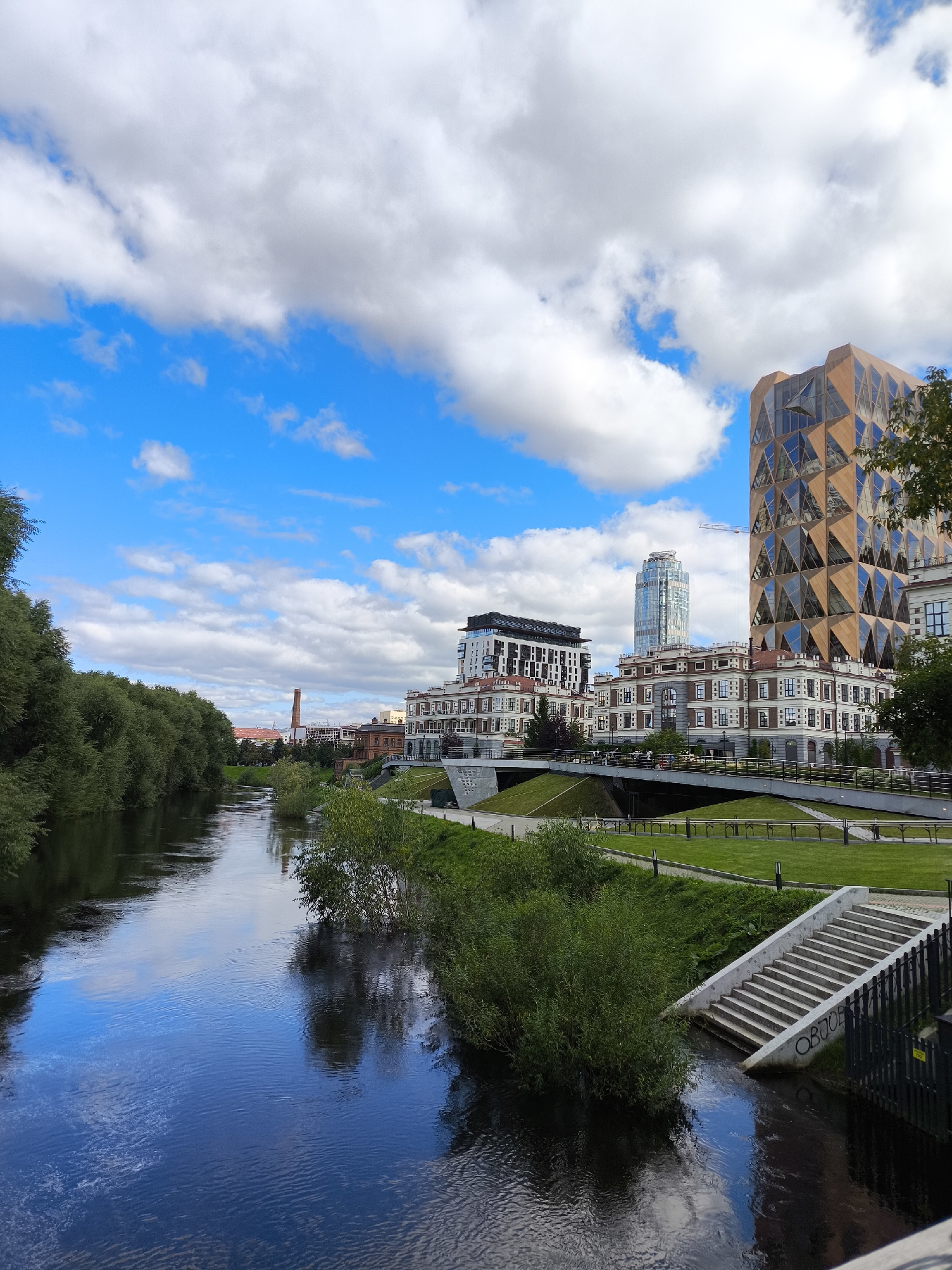 The image shows the Iset River in Yekaterinburg, Russia, with modern buildings along its banks. On the right side of the river, several contemporary buildings are visible, including a striking tower with a geometric, faceted facade in shades of wood and glass. Adjacent to it are lower buildings with a classic architectural style, featuring red brick and white trim. A tall, slender skyscraper with a glass facade stands further in the background, and a construction crane is visible behind it. On the left side of the river, a dense line of green trees forms a natural border. The river itself is dark and reflects the blue sky and white clouds, with ripples on its surface. A concrete staircase descends from a grassy embankment towards the river. The sky is a vibrant blue, dotted with scattered white clouds. The overall scene depicts an urban landscape where nature and modern architecture coexist.