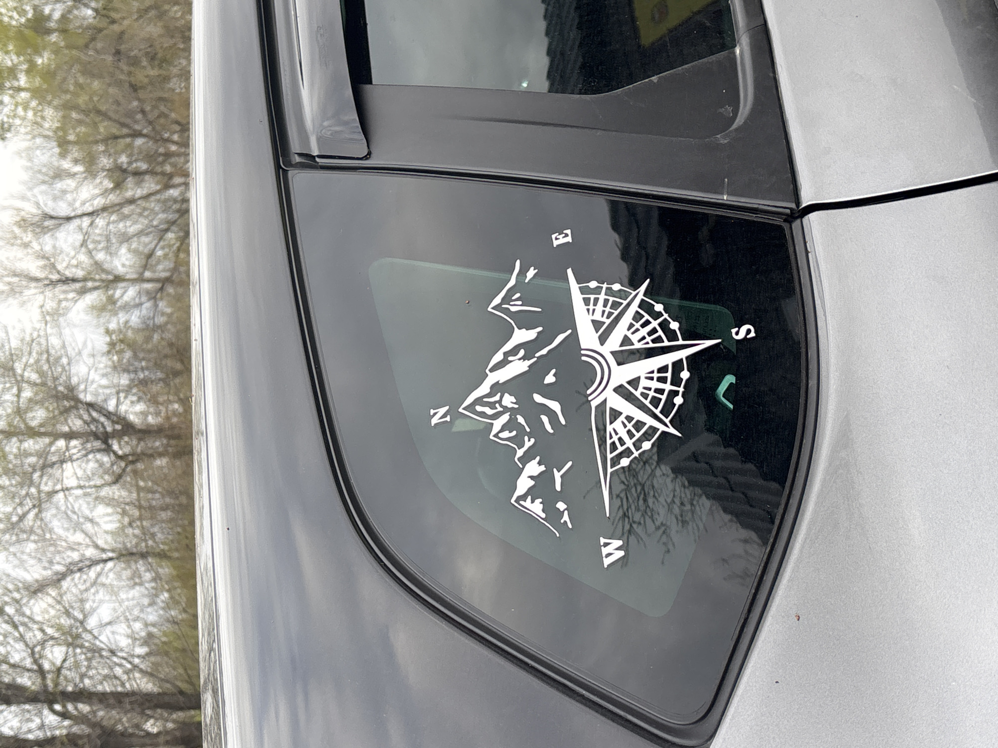 The image displays the rear passenger window and surrounding bodywork of a grey car, viewed from the side. The window is adorned with a white vinyl decal depicting a stylized mountain range integrated with a compass rose.
The decal features a jagged, multi-peaked mountain silhouette on the left, conveying an adventurous or outdoor theme. To the right, a detailed compass rose clearly marks the cardinal directions with the letters "N" (North) at the top, "E" (East) to the right, "S" (South) at the bottom, and "W" (West) to the left. The compass rose itself has an intricate design with multiple points and internal grids.
The car's body is a light grey, while the window trim and B-pillar are black. Reflections in the car window show the bare or sparsely leafed branches of numerous trees against a bright, overcast sky. This suggests an outdoor setting during a cooler season, possibly late winter or early spring. The car is stationary, and no individuals are visible. Small specks of dust or dirt are noticeable on the car's paint and window. The lighting is diffused, consistent with an overcast daytime. The scene is situated in Orenburg, Russia.