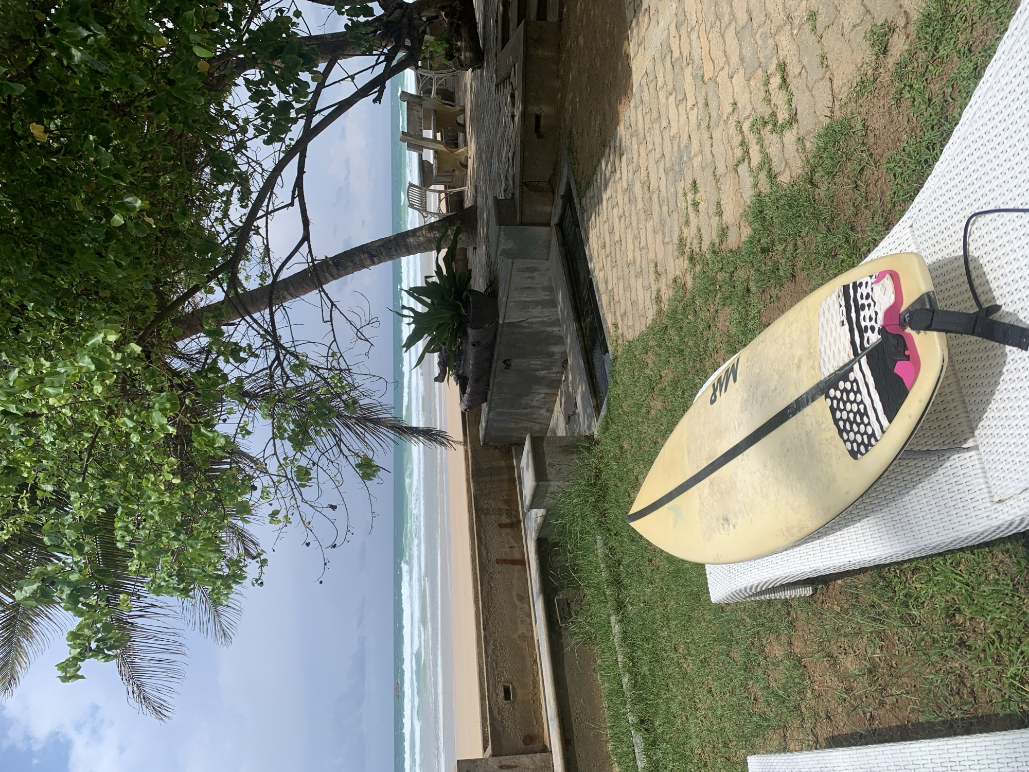 A rotated medium shot captures a sunny beach scene in Kathaluwa, Sri Lanka. A tan surfboard with black and white designs rests on a white wicker chaise lounge, positioned on a lawn alongside a brick-paved area. A low concrete wall divides the lawn from the sandy beach. Beyond the wall, gentle waves roll onto the shore. A cluster of chairs and tables sit under trees providing shade. Lush tropical foliage frames the scene, partly obscuring the sky.