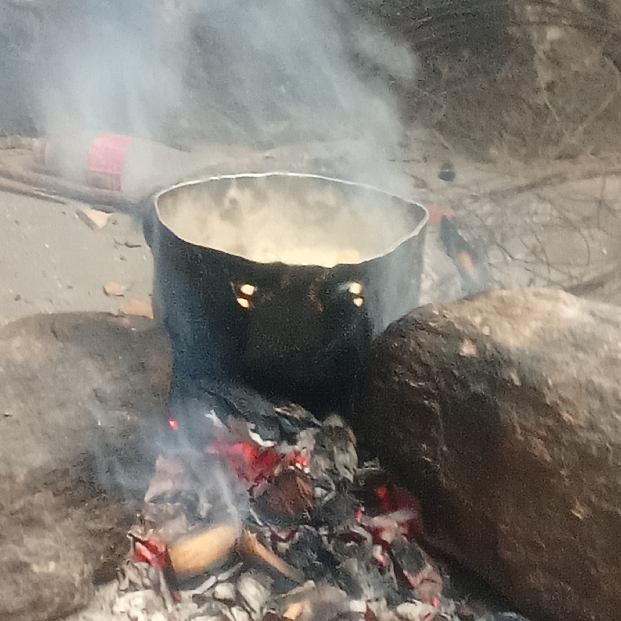 A black cooking pot sits atop an open fire, supported by rocks on either side. Smoke rises from the fire and the pot. A red and white bottle is visible in the background to the left of the pot. The surrounding environment appears to be a natural, outdoor setting. The location is Unknown, Unknown.