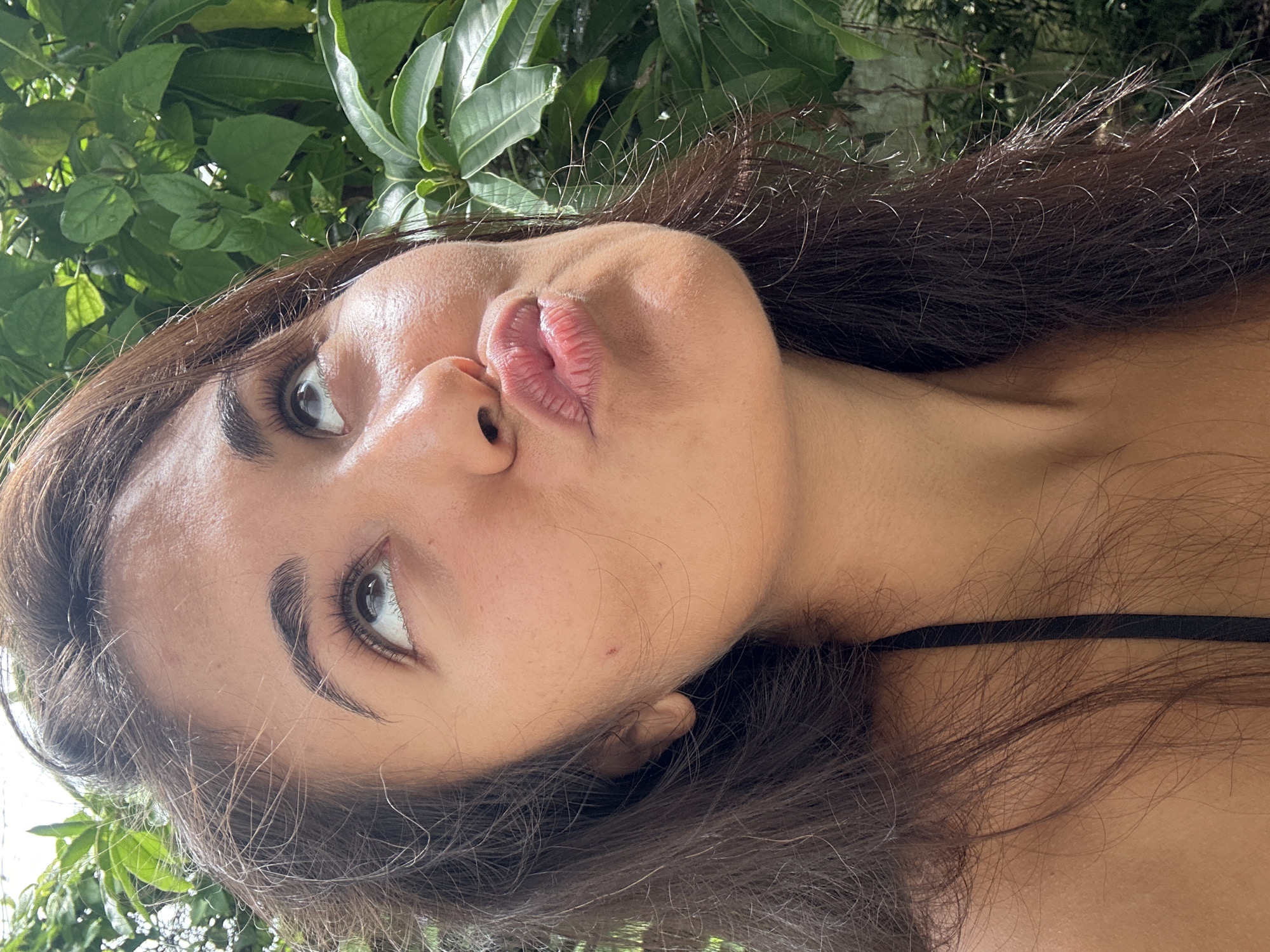 A close-up, sideways selfie of a young woman with long, dark hair is captured against a backdrop of lush green foliage, suggesting an outdoor setting in a tropical location like Peleana, Sri Lanka. The woman's face is partially visible, with her eyes looking to the left and her lips pursed in a playful pout. Her skin appears smooth and sun-kissed, with a few faint freckles visible on her cheek. Her dark eyebrows are neatly shaped, and her eyelashes are long and full, framing her expressive eyes. Her lips are full and slightly parted, with a subtle sheen that suggests lip balm or gloss. The lighting appears natural, likely from daylight, casting gentle shadows and highlighting the textures of her skin and hair. The leaves in the background are vibrant and detailed, indicating a bright, possibly sunny, day. The overall mood is casual and relaxed, with a hint of flirtatiousness conveyed through her pose and facial expression. There is no visible text in the image.