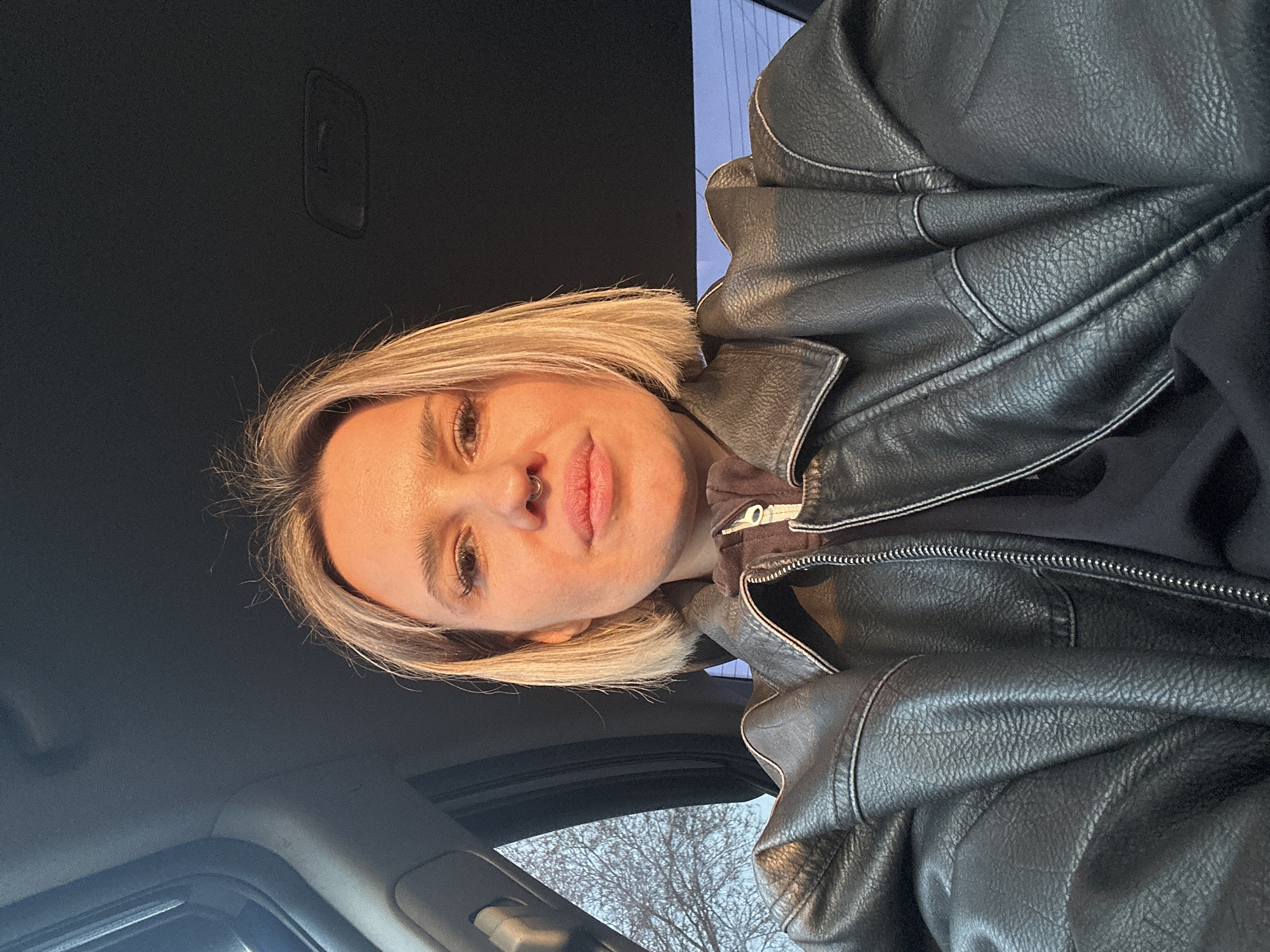 An adult female is depicted in a close-up, chest-up shot, seated within the interior of a vehicle. She has short, straight, blonde hair with visible roots, and a silver-colored septum piercing. Her facial expression is neutral, and she is looking directly at the camera. She is wearing a black, leather-textured jacket featuring light-colored stitching on the seams, over a dark, possibly hooded, garment with a partially visible silver zipper. Warm, directional light, consistent with late afternoon or early evening sunlight, illuminates her face from the left, casting an orange hue on her skin. Above her head, the dark gray or black interior headliner of the vehicle is visible. To her left, through the vehicle's window, a bare tree with thin, leafless branches is visible against a light blue-grey sky. There are no identifiable landmarks or specific geographical features within the image to confirm the location as Rostov-on-Don, Russia.