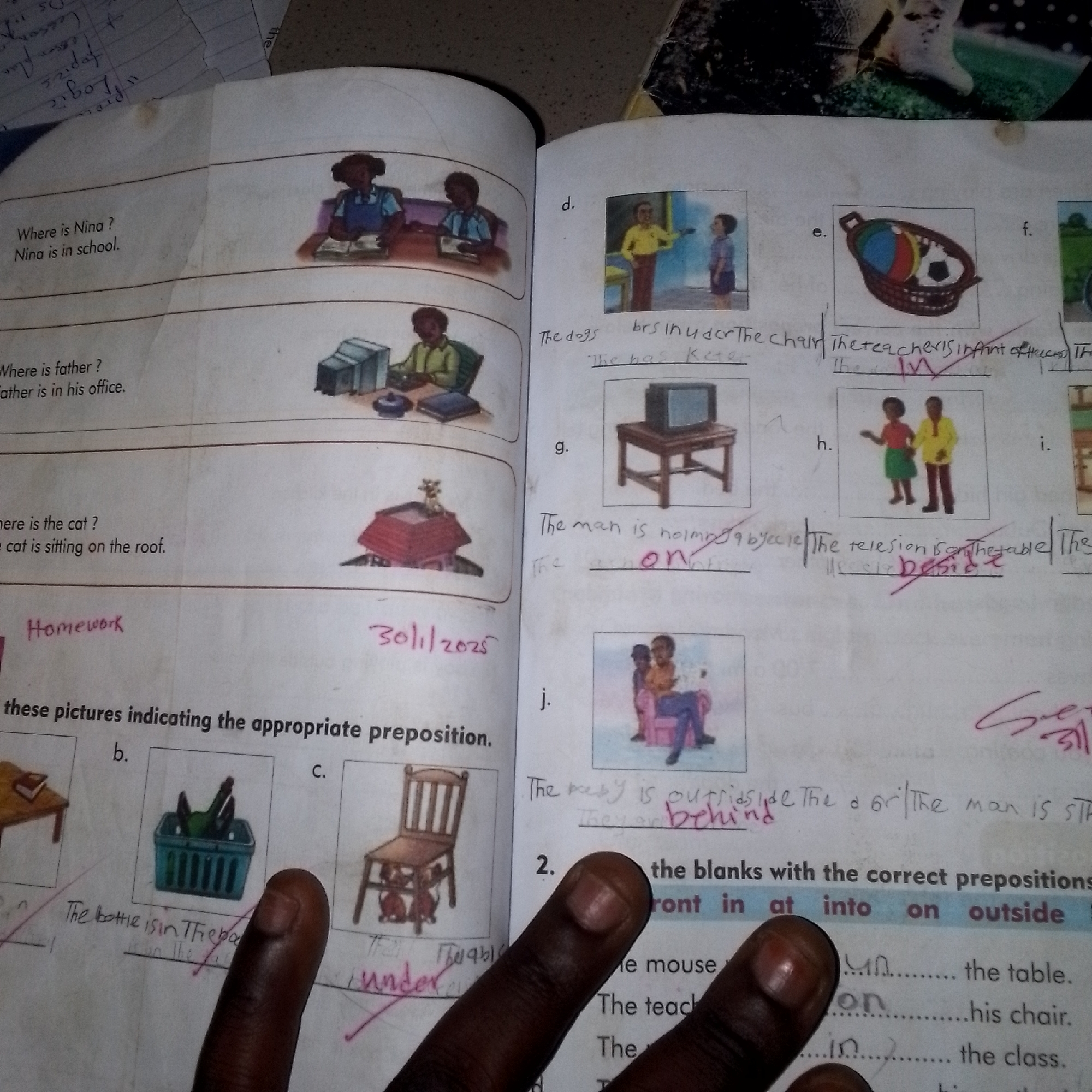 This is a close-up, overhead shot of an open workbook or textbook. The page is primarily white with printed text and illustrations, some of which have been colored in. There is also handwritten text and markings on the page.
On the left side of the page, there are three questions and corresponding images.
The first question asks "Where is Nina?" with the answer "Nina is in school." This is accompanied by an illustration of a girl and a boy sitting at desks in a classroom.
The second question asks "Where is father?" with the answer "Father is in his office." This is accompanied by an illustration of a man sitting at a desk with a computer.
The third question asks "Where is the cat?" with the answer "The cat is sitting on the roof." This is accompanied by an illustration of a cat on a roof.
The right side of the page contains several numbered sections (d, e, f, g, h, i, j) with smaller illustrations and accompanying text, some of which has been filled in or corrected by hand.
Section d shows two boys.
Section e shows a basket with items in it.
Section g shows a television on a stand.
Section h shows two people.
Section j shows two people sitting on a sofa.
Below the illustrations, there is a section titled "these pictures indicating the appropriate preposition." There are also two numbered sections with fill-in-the-blank sentences.
The bottom of the page has a section with a list of prepositions: "ont in at into on outside".
There are several instances of handwritten text, including "Homework", a date "30/11/2025", and various answers or corrections to the exercises. A finger is visible in the foreground, obscuring parts of the lower section of the page. The overall impression is of a school assignment or workbook being used.
There is no information in the image to suggest the location of Jalingo, Nigeria.