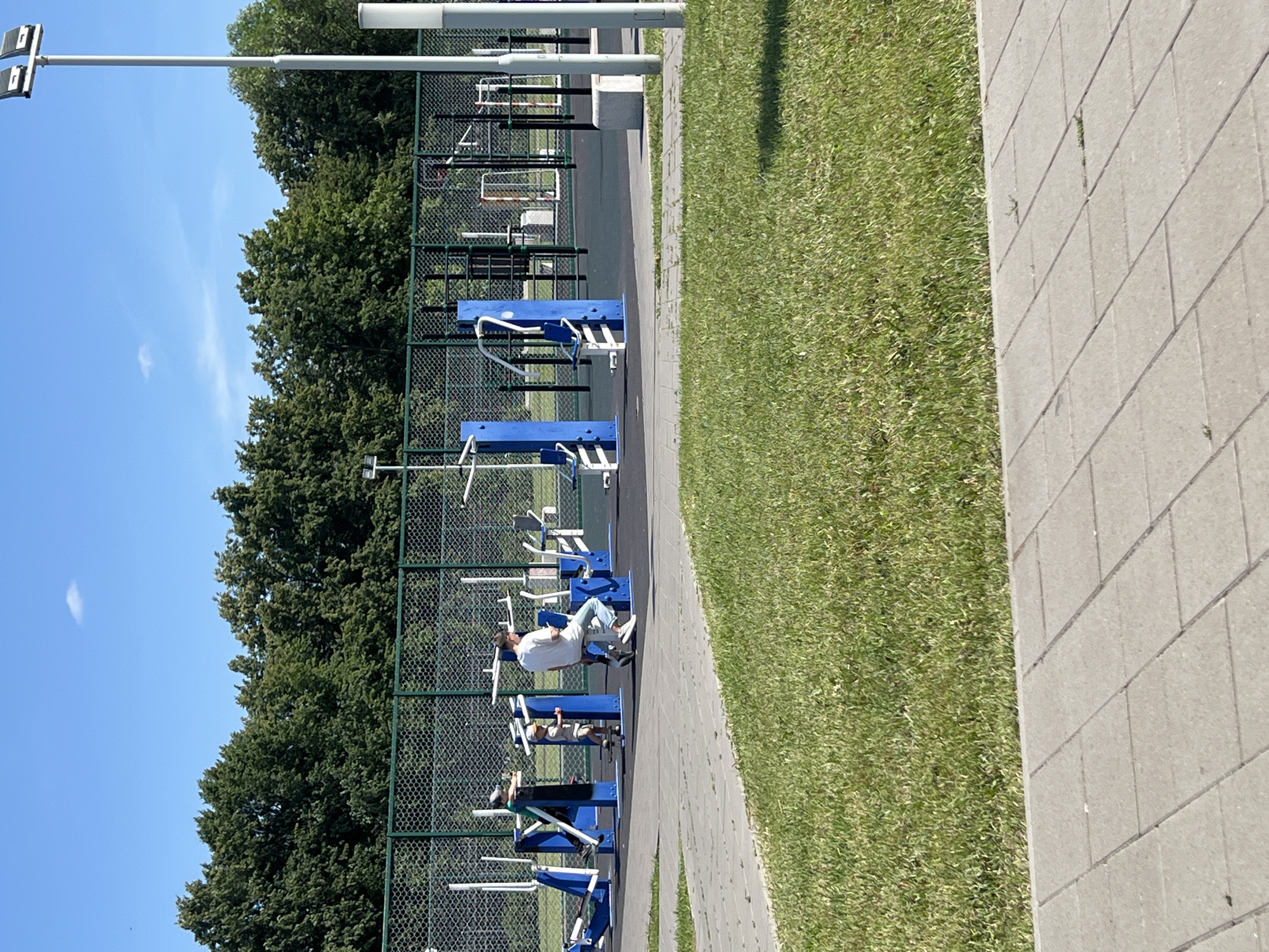 This media file depicts an outdoor gym scene at a park or recreational area, likely in Tolyatti, Russia. The foreground features a paved walkway bordering a well-maintained grassy lawn. Beyond the walkway, a chain-link fence encloses a series of blue and white outdoor fitness equipment. One person, dressed in a white t-shirt and jeans, is actively using a weight machine, positioned on the equipment and appearing engaged in their workout. Another person, wearing a grey shirt and shorts, is visible on a different piece of equipment, also seemingly exercising.
The setting is bathed in bright daylight, suggesting it's daytime. The sky is clear and blue with a wispy white cloud in the distance. Lush green trees form a dense backdrop behind the gym equipment and fence. The overall impression is one of a pleasant, active day in a public outdoor space designed for physical fitness. The equipment appears to be designed for a variety of exercises, with visible handles, weights, and seating. The scene is peaceful and functional, focused on exercise and recreation. No visible text is present in the image.