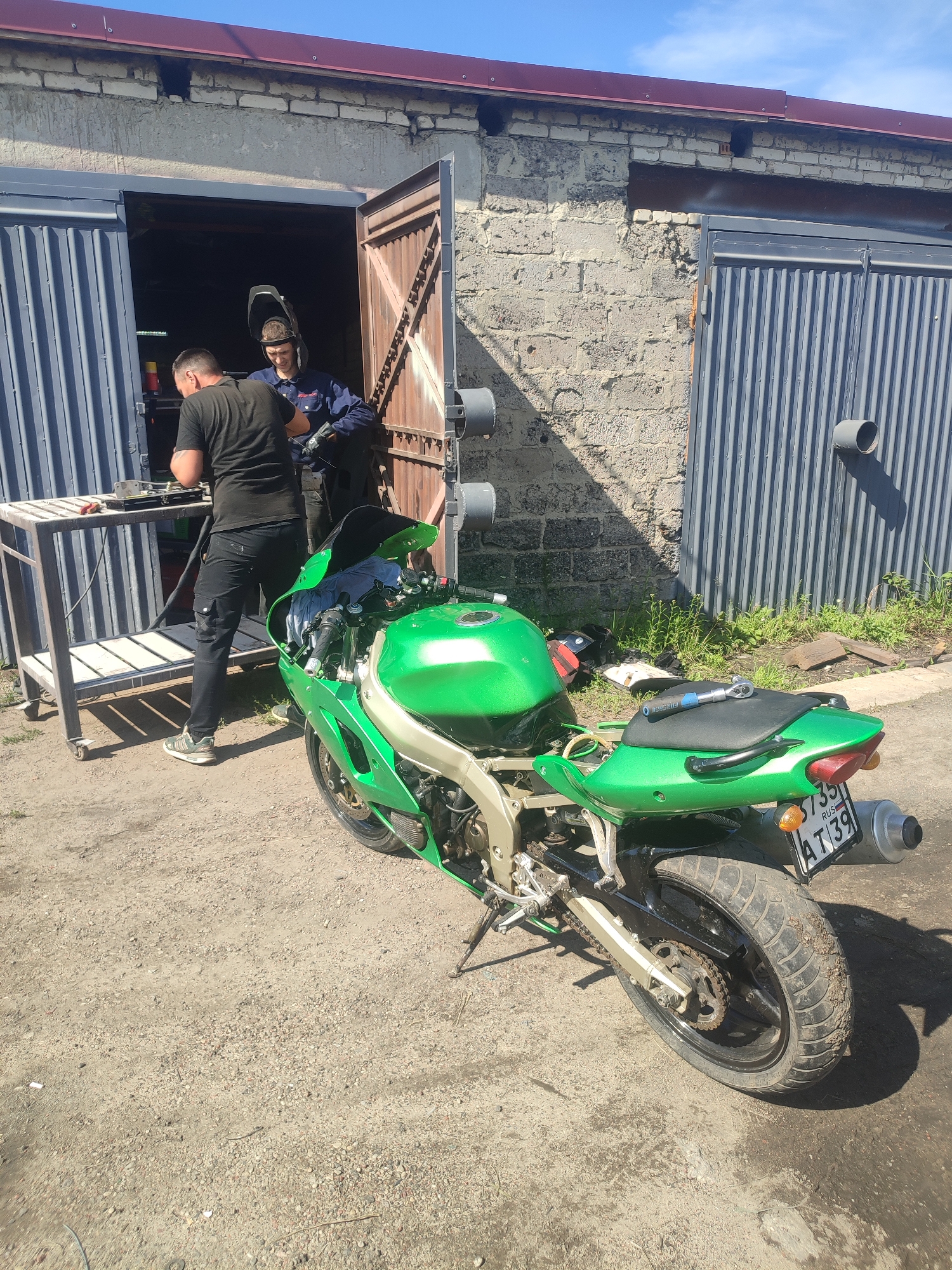 Two individuals are present outside a garage with a green motorcycle. One person, wearing a black t-shirt and pants, stands near a metal workbench. The second person, wearing a blue uniform and welding helmet, is inside the garage doorway. The motorcycle is partially disassembled and parked on a gravel surface. The garage appears to be an industrial or workshop setting, possibly in Zelenogradsk, Russia, based on the motorcycle's license plate which reads "3735 AT39".