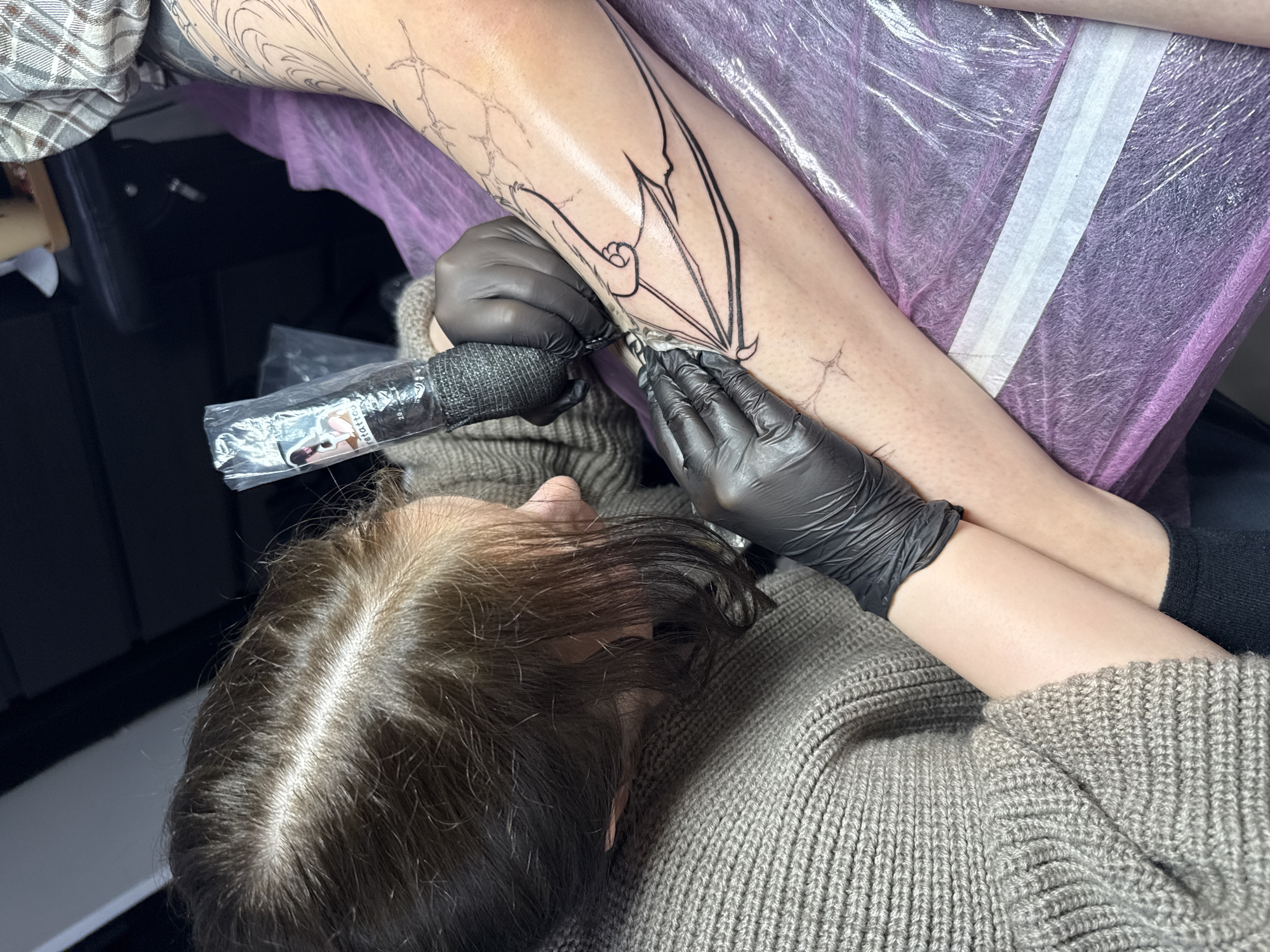 A person with fair skin is receiving a tattoo on their left leg, likely the calf or lower leg. The tattoo is a black outline of a bat-like design. The tattoo artist, wearing black gloves and a gray knitted sweater, is holding a tattoo machine, which is wrapped with a black grip. The artist's hair is dark and pulled back. The leg is on a surface covered with a purple and white plastic sheet. A plastic bag with tattoo-related items inside is visible near the tattoo machine. The scene suggests an indoor environment, possibly a tattoo parlor. The city is Unknown, Unknown.
