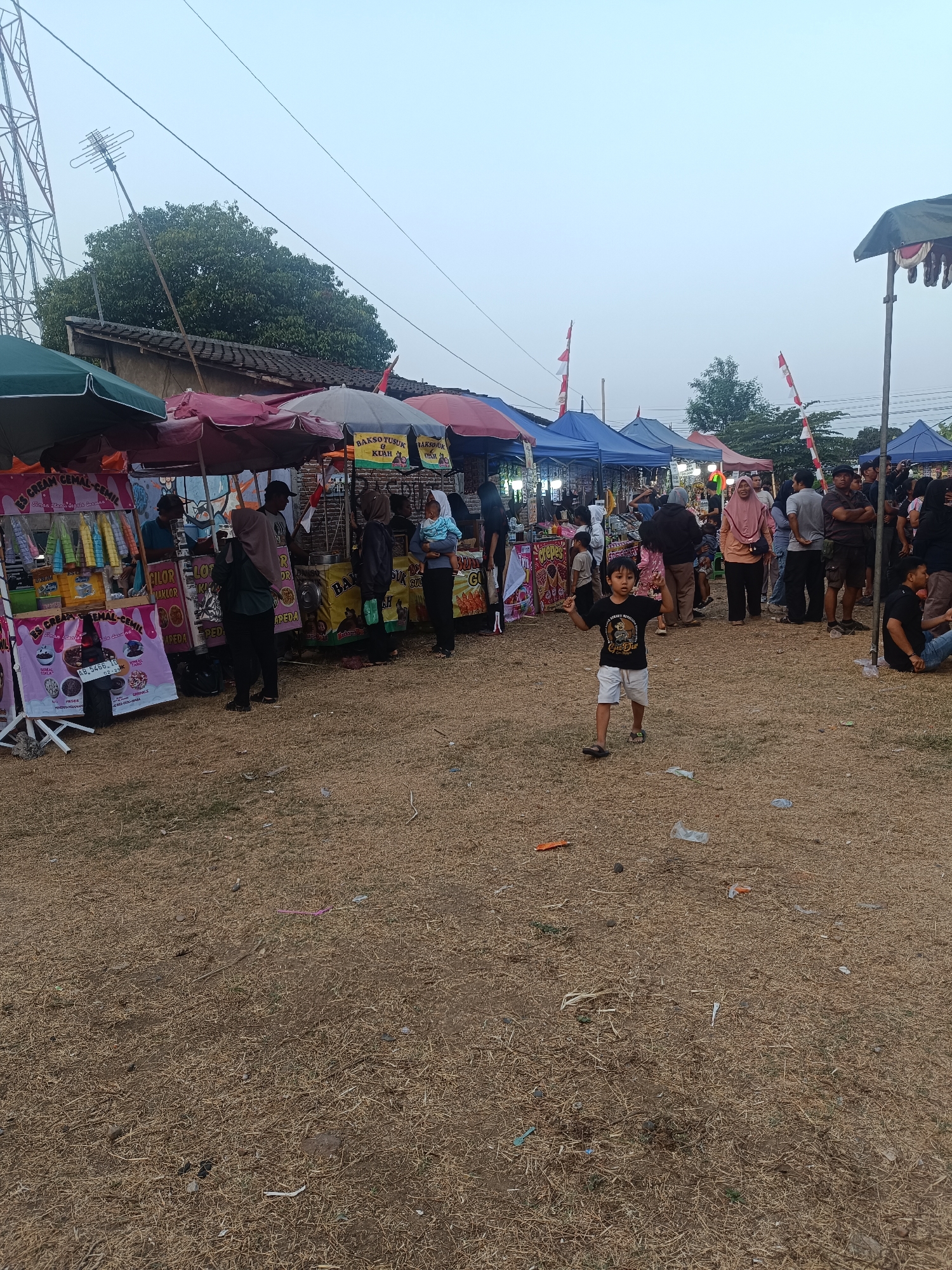 The image depicts an outdoor market scene in Jetis, Indonesia, possibly during a festival or local event. In the foreground, a young boy wearing a black t-shirt with graphics and white shorts is captured mid-stride, his arms raised. The ground is covered with dry, sparse grass and scattered debris, including plastic wrappers and small pieces of trash.
Behind the boy, a row of market stalls with colorful awnings are set up. Several people are seen browsing or working at these stalls. Some stalls display food items like "Bakso Tusuk" (skewered meatballs) and "Es Cream Cemal-Cemil" (snack ice cream), indicated by signage. Tents in various colors, including blue and red, form the backdrop of the market. Indonesian flags are visible on poles above some of the stalls, suggesting a national celebration or event.
In the mid-ground, a dense collection of people are gathered, some appearing to be customers and others vendors. Beyond the stalls, a large green tree stands tall, and a building with a tiled roof is partially visible. To the far left, a tall telecommunications tower with an antenna is a prominent feature of the landscape. The sky is overcast and a pale blue, indicating either early morning or late afternoon light. The overall atmosphere suggests a bustling local gathering.