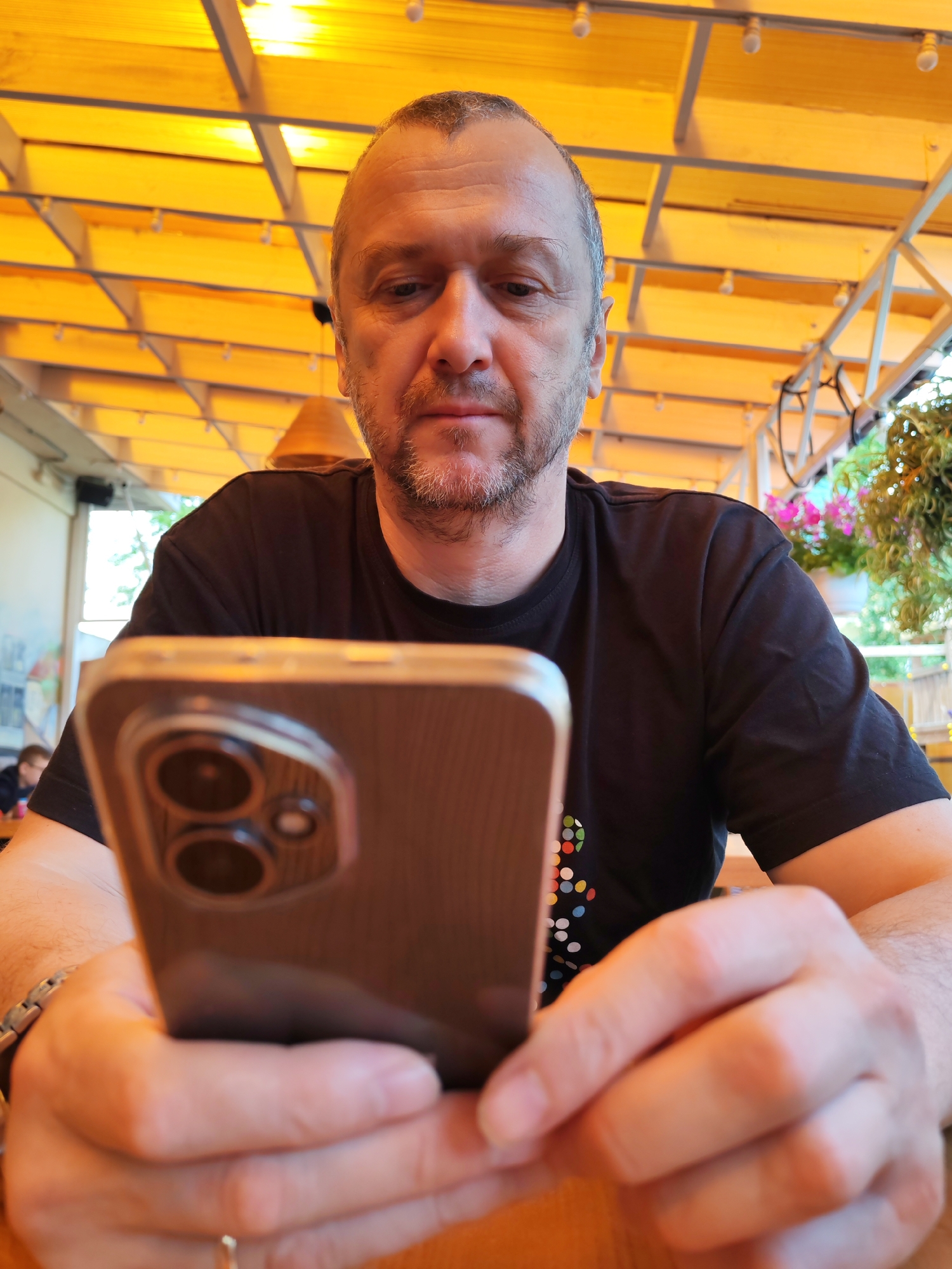 The image shows a man with short brown hair and stubble looking down at a smartphone held in his hands. He is wearing a black t-shirt. The phone has a clear case and a triple-lens camera on the back. The man is seated at a wooden table, and the background suggests an outdoor or covered seating area, possibly a cafe or restaurant, with a yellow canopy overhead. In the background, there are hanging plants with pink flowers and other patrons. The overall impression is a casual setting. This scene is located in the village of Bol'shie Mkhi, Russia.
