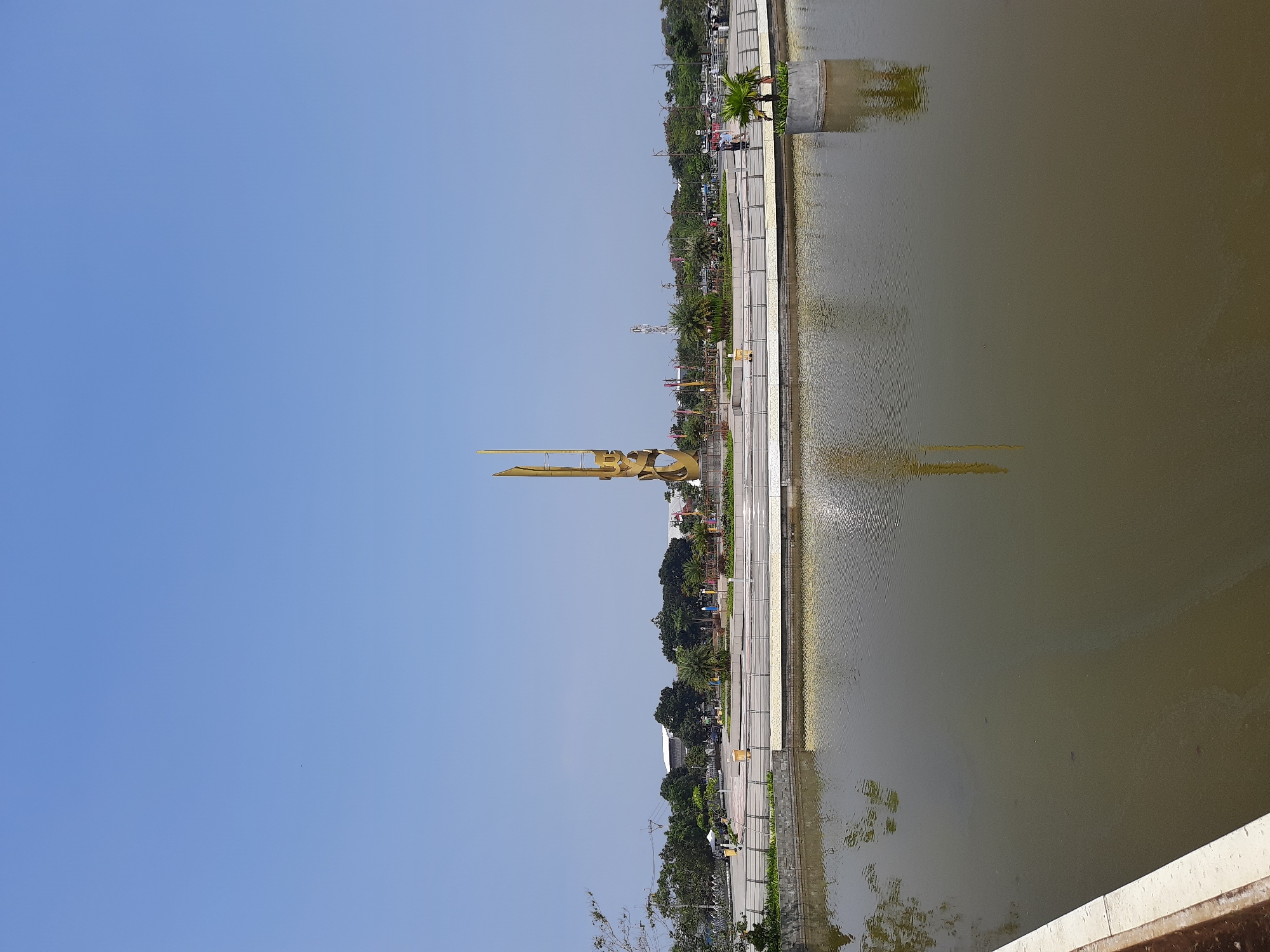 This is a daytime, wide-angle, eye-level shot of a city park with a large pond in the foreground and a modern monument in the background. The pond's water is murky brown and reflects the clear blue sky and the golden monument. A white-tiled ledge lines the bottom left of the pond. The middle ground features a paved walkway surrounding the pond, bordered by a metal railing. Beyond the walkway, a raised circular area with greenery and some palm trees is visible. The dominant feature in the background is a tall, abstract golden monument with intricate geometric designs. Surrounding the monument is a park-like setting with scattered trees, buildings, and a few people walking. The sky is clear blue, with no clouds. The overall scene suggests a public gathering or recreational space in an urban environment. This scene appears to be the Alun-Alun Kota Bandung, a public square in Bandung City, Indonesia, featuring the Monumen Bandung Lautan Api.
