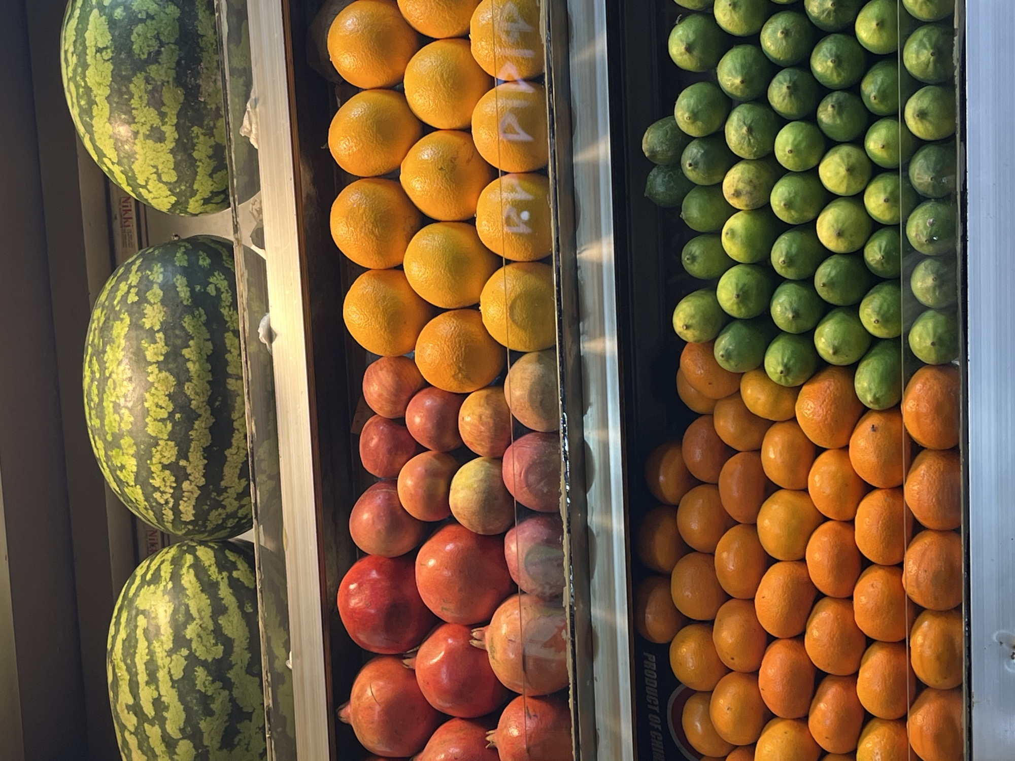The image shows a display of fruit in what appears to be a shop or market in Dhaka, Bangladesh. The fruit is arranged in vertical columns: watermelons, oranges, apples, pomegranates, limes, and more oranges. The display is viewed through a glass window or partition, and there's writing on the oranges. The produce is densely packed, creating a colorful and organized arrangement.