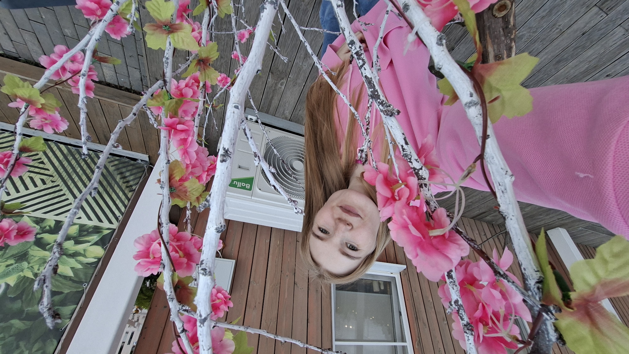 A young woman with long, straight brown hair and light eyes is pictured in an outdoor setting in Chaikovsky, Russia. She is wearing a bright pink long-sleeved top and is looking directly at the camera with a neutral expression. The image is taken from a low angle, with the woman's head and upper torso visible as she looks down.
The foreground is dominated by white, branch-like structures adorned with artificial pink flowers and light green leaves, creating a decorative frame around the woman. In the background, there is a wooden-sided building, suggesting a rural or suburban environment. An air conditioning unit is visible on the side of the building, with the brand name "Ballu" clearly readable on its front panel. A window with a white frame is also partially visible on the building. To the left, there's a banner or backdrop with green foliage and a black and white geometric pattern.
The lighting suggests it is daytime. The overall impression is one of a posed portrait taken outdoors, possibly in a garden or on a porch decorated with artificial floral arrangements.