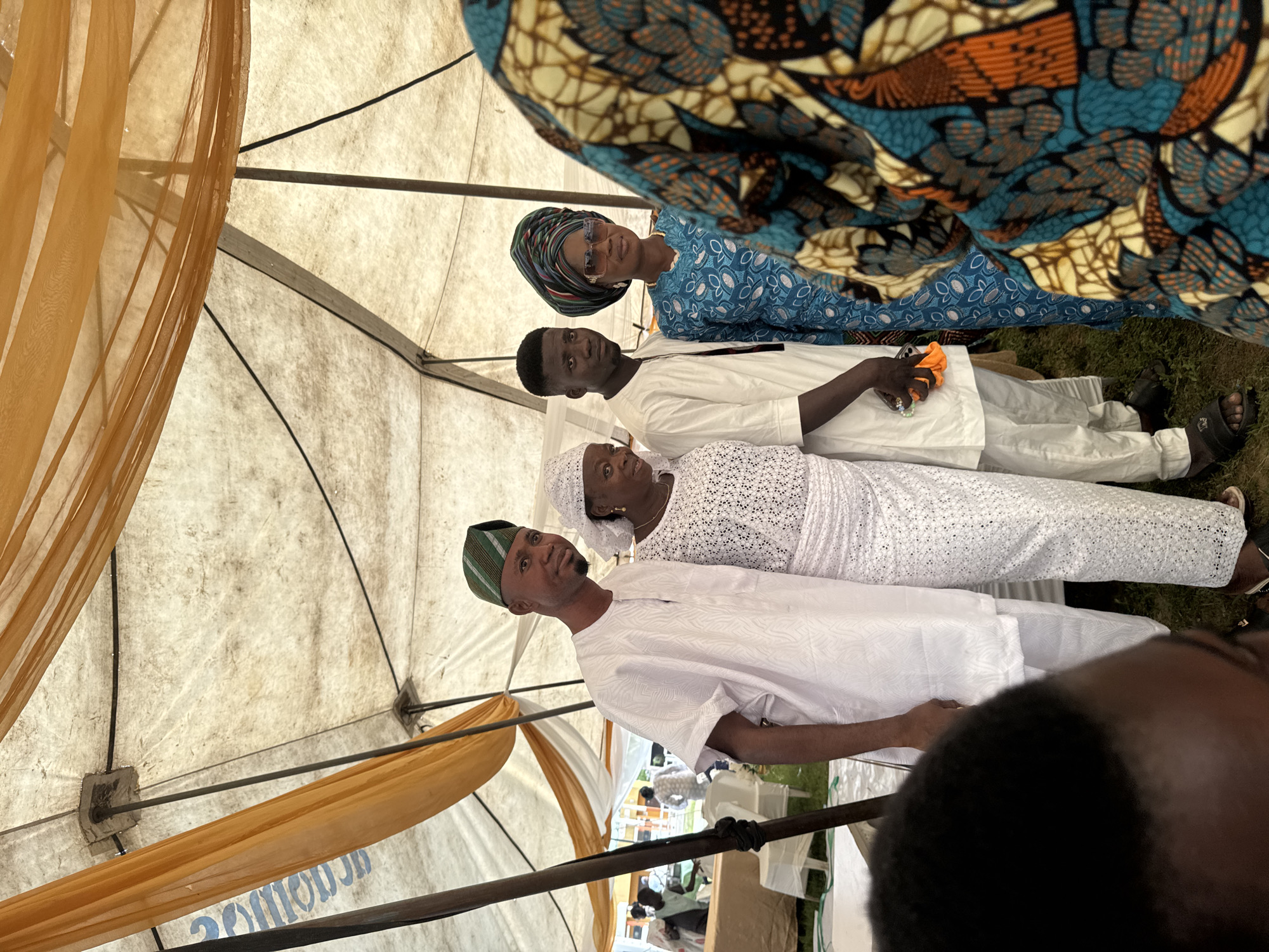 In an outdoor event under a white tent with orange draped fabric, four individuals are posed for a photograph. A man on the left wears a white traditional outfit and a green and striped cap. Beside him, a woman is dressed in a white lacy outfit and head covering. To her right, a young man in a white traditional outfit stands next to a woman wearing a blue patterned dress and a colorful headwrap, adorned with sunglasses. The background shows blurred figures and white chairs, suggesting a gathering or celebration in Badagry, Nigeria.
