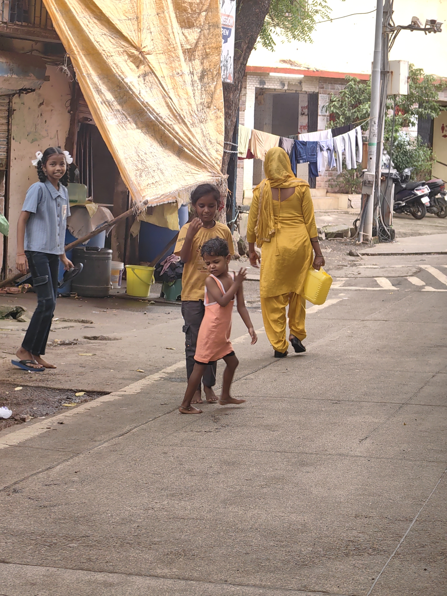 A wide shot captures an outdoor scene in Mira-Bhayander, India, with four people and various objects on a paved surface. In the foreground, a young girl in an orange dress with white straps is captured mid-stride, her arms extended outwards, possibly playing. To her left, a slightly older girl in a yellow t-shirt and dark pants stands and looks towards the foreground. Behind her, another young girl in a light blue collared shirt and dark pants walks away from the camera, holding a dark blue flip-flop in her right hand. In the background, a woman in a bright yellow outfit, including a headscarf, walks towards the right, carrying a yellow plastic jerrycan in her right hand. Her back is to the camera.
The environment includes a weathered, yellowish tarp stretched across the top left of the frame, supported by poles. To the left of the tarp, a dilapidated building with peeling paint and an open doorway is visible. Several large dark grey cylindrical containers and a bright yellow bucket are situated near the building. In the mid-ground, laundry is hung on a line between a tree and a pole, with more trees and a building visible in the distance. A portion of a street with white lines marking a crosswalk is also present. A dark-colored scooter is parked on the right side of the frame, partially obscured by a utility pole. The ground is a textured, grey paved surface, with scattered debris and puddles of water in some areas. The lighting suggests it is daytime with overcast conditions.