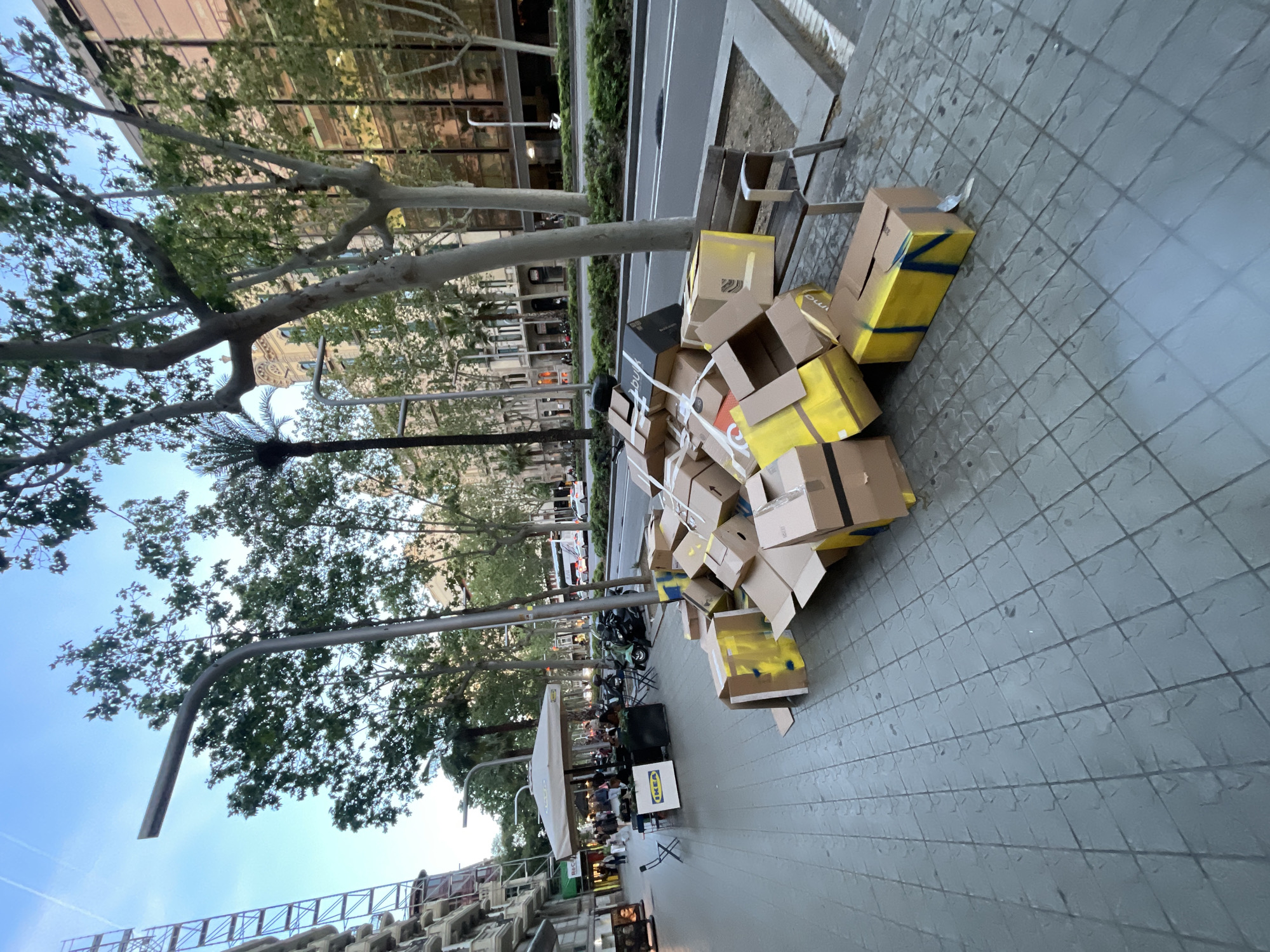 This urban street scene, oriented vertically, depicts a bustling sidewalk alongside a road in what appears to be late afternoon or early evening. The main subject is a large, untidy pile of cardboard boxes stacked on a grey tiled pavement in the foreground and midground. Many of these boxes are standard brown cardboard, while several are partially painted yellow or secured with yellow tape. Some black boxes are also mixed into the pile.
To the left of the box pile, a street bench is visible. Further down the sidewalk, an outdoor seating area of a cafe or restaurant is set up under a white canopy. Several people are seated at tables in this area. A prominent white sign with the blue and yellow "IKEA" logo stands near the outdoor seating.
Beyond the sidewalk, a road with at least two lanes is visible, bordered by a narrow median strip with green bushes. Parked motorbikes and other vehicles can be seen along the street. Large, mature trees with green foliage line the street, casting some shadows. In the background, modern buildings with reflective windows and classical architectural elements are visible, suggesting a commercial or residential urban area. Street lamps with curved arms extend over the sidewalk. The sky is clear and blue.
The primary activity shown is people relaxing at an outdoor cafe. The pile of boxes suggests either a delivery point, discarded packaging, or items awaiting recycling. Notable text includes "IKEA" on a sign and a box, and "bulub" on a black box, and "RAC" on a green box. The lighting indicates a clear day, likely late afternoon or early evening. The scene is consistent with an urban setting, specifically Barcelona, Spain, given the provided context.