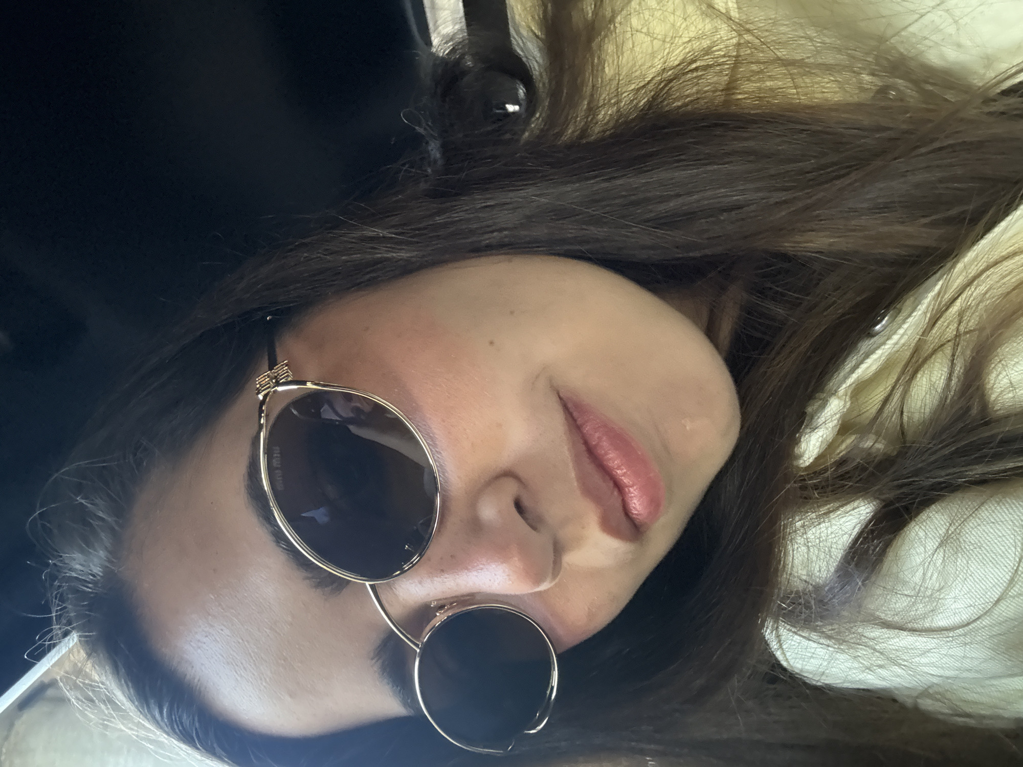A woman with dark hair and sunglasses is seen in an indoor setting. The frame of the sunglasses is golden, and the lenses are dark. The woman's skin appears light, and her lips are subtly pink. The background includes dark areas and lighter, blurry patches of what could be clothing. The scene appears to be in a car, but there is no indication that it is in Mexico City.