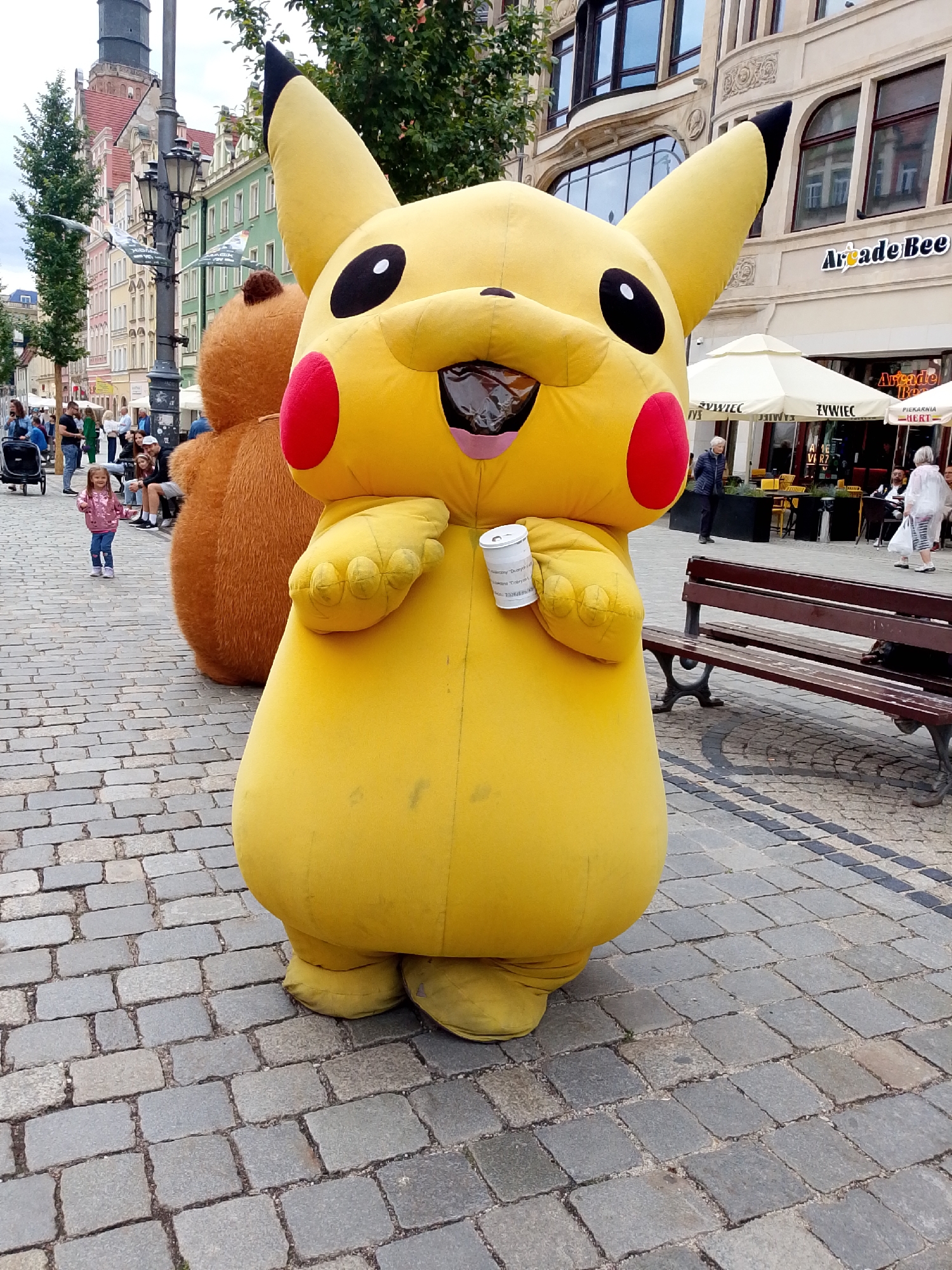 The image shows a Pikachu mascot costume standing on a cobblestone street in Wrocław, Poland. The mascot is large and yellow with red cheeks and black eyes. It is holding a white cup in its right hand. Behind the Pikachu, a large brown teddy bear mascot is visible. To the left of the mascots, a young girl in a pink jacket is walking on the cobblestone street. In the background, there are buildings with windows and signs, as well as people sitting at outdoor tables and walking. There is also a park bench on the right side of the image. The environment suggests a public outdoor space in a European city, likely a pedestrian zone or a town square. The buildings in the background appear to be historic. The sky is overcast.