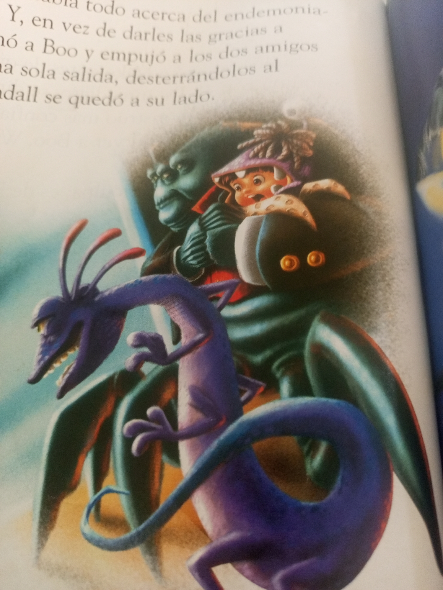 The image is a close-up photograph of an open book, showing an illustration and text. The illustration features several animated characters, primarily from the movie "Monsters, Inc." In the foreground, a purple, lizard-like creature with a long tail and sharp teeth is prominent, appearing to be running. Behind it, a larger, dark-skinned creature with green skin and large, round eyes is visible. A small child, likely Boo from the movie, is depicted wearing a monster costume with a purple hat. The text on the left side of the page is in Spanish and discusses giving thanks, implying a narrative context. There is no indication that this image is from La Tagua, Venezuela.