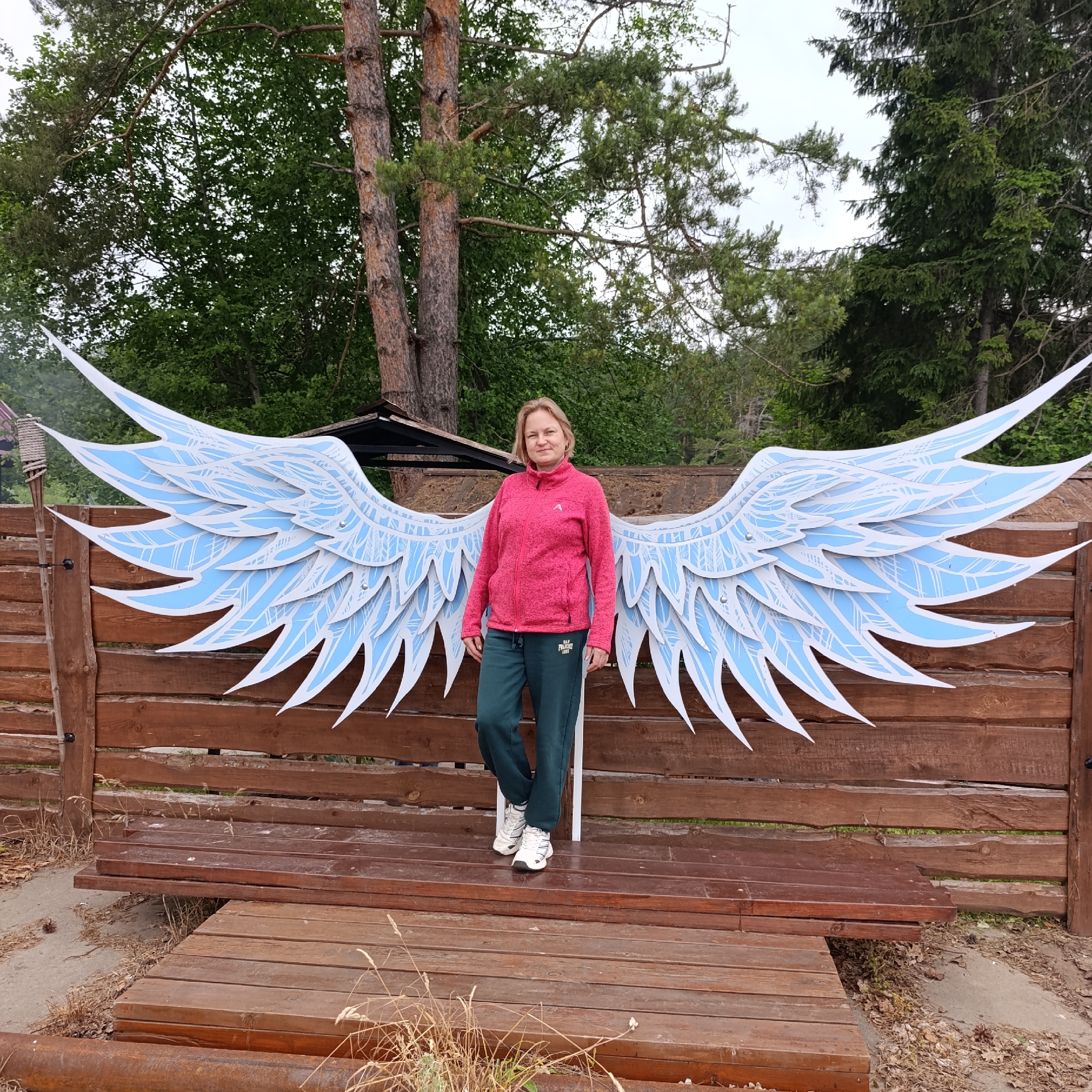 In Prikamskii, Russia, a Caucasian woman with blonde hair stands in front of a large, light blue and white, stylized angel wing decoration. She is wearing a pink zip-up jacket and dark green sweatpants, along with white athletic shoes. The wings are mounted on a rustic wooden fence, and the woman is positioned on a wooden platform in front of it. The background consists of lush green trees and a cloudy sky. The scene appears to be outdoors, possibly in a park or recreational area, given the natural setting and the decorative installation.