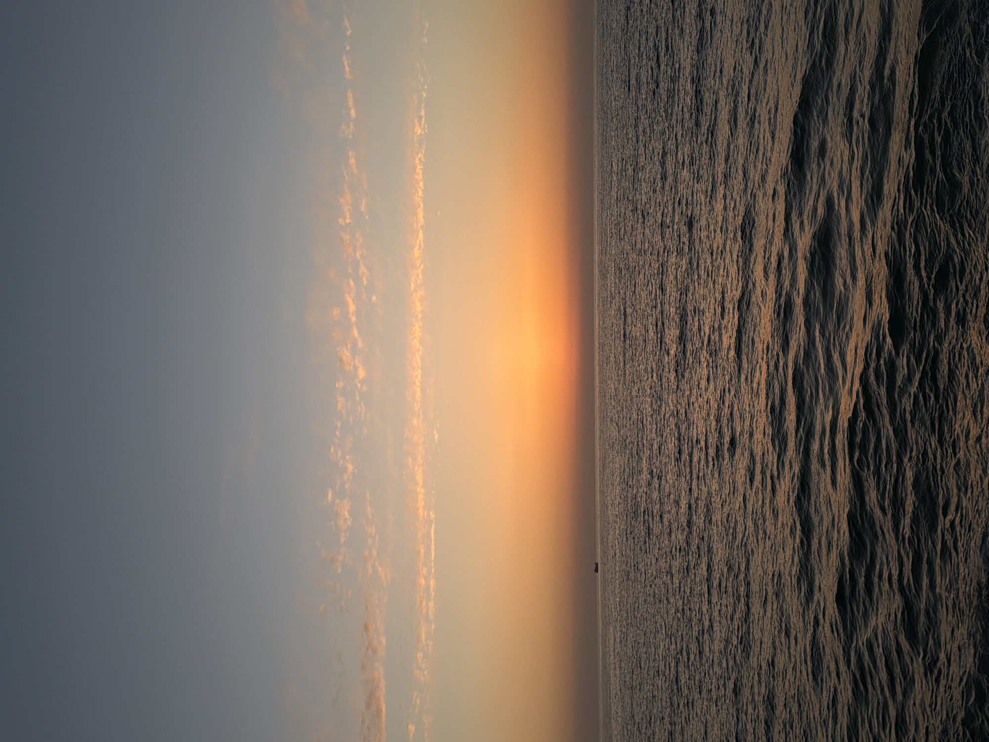 The image captures a sunset over the Mediterranean Sea in Tel Aviv, Israel. The sky transitions from blue to orange, with long, thin clouds catching the light. The sea, slightly choppy, reflects the sunset colors. A small boat is visible on the horizon. The image is taken from a high vantage point, likely along the Tel Aviv coast.