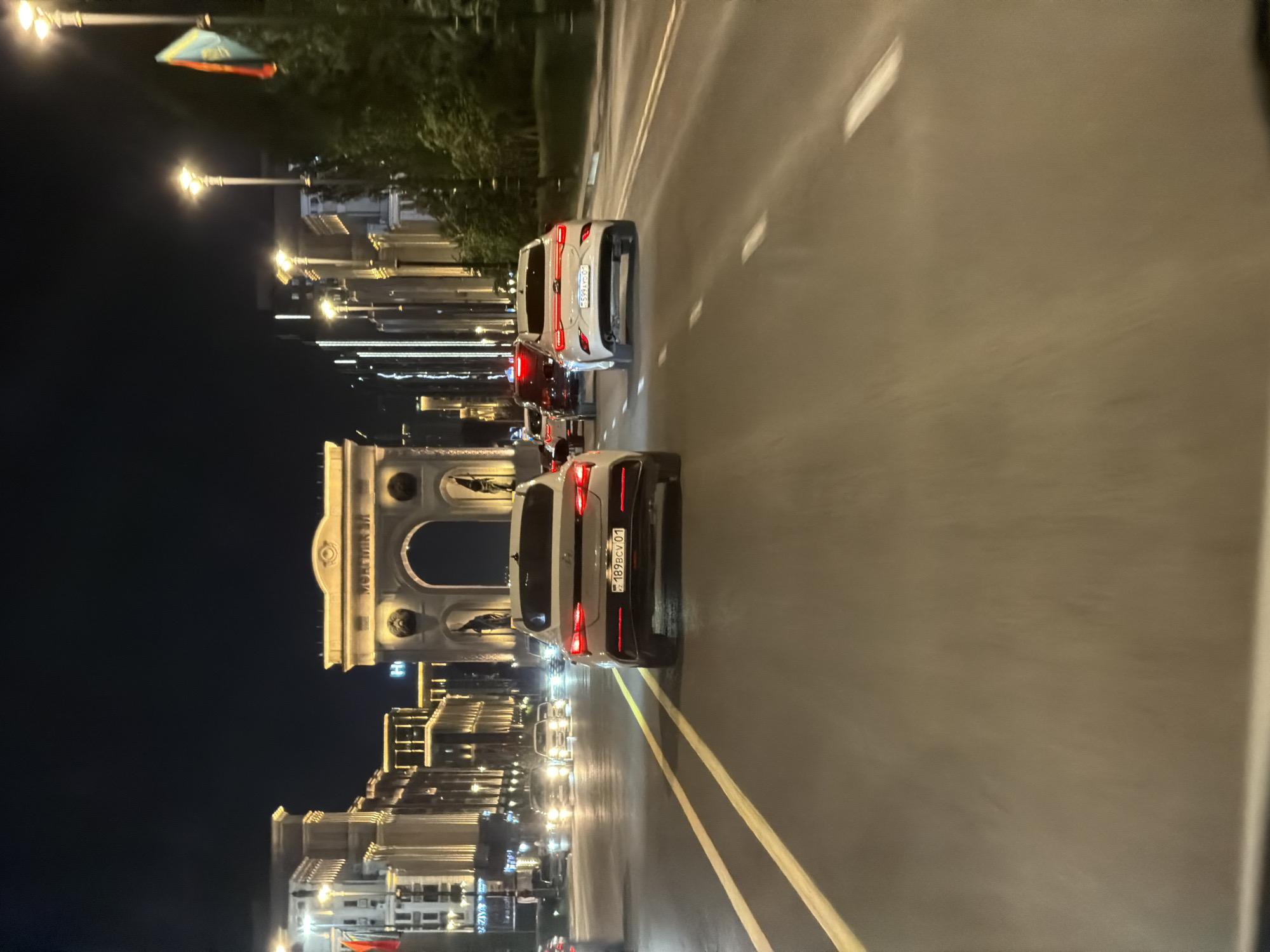 The image captures a nighttime view of a city street in Astana, Kazakhstan, featuring several vehicles driving on a multi-lane road. In the background, a large archway, lit with golden lights, stands prominently. Adjacent to the archway are illuminated buildings with ornate architectural details, suggesting a significant urban landmark or historic district. Streetlights also dot the scene, casting a warm glow on the surroundings. Several cars are visible on the road, including a white sedan in the foreground with its license plate reading "189BCV 01". Another white car is ahead of it, and a red car is further down the road. The road surface is asphalt, marked with yellow lane lines. Trees and foliage can be seen on the right side of the image, partially obscuring the view of some of the background elements. The overall impression is one of a vibrant, well-lit urban environment at night.