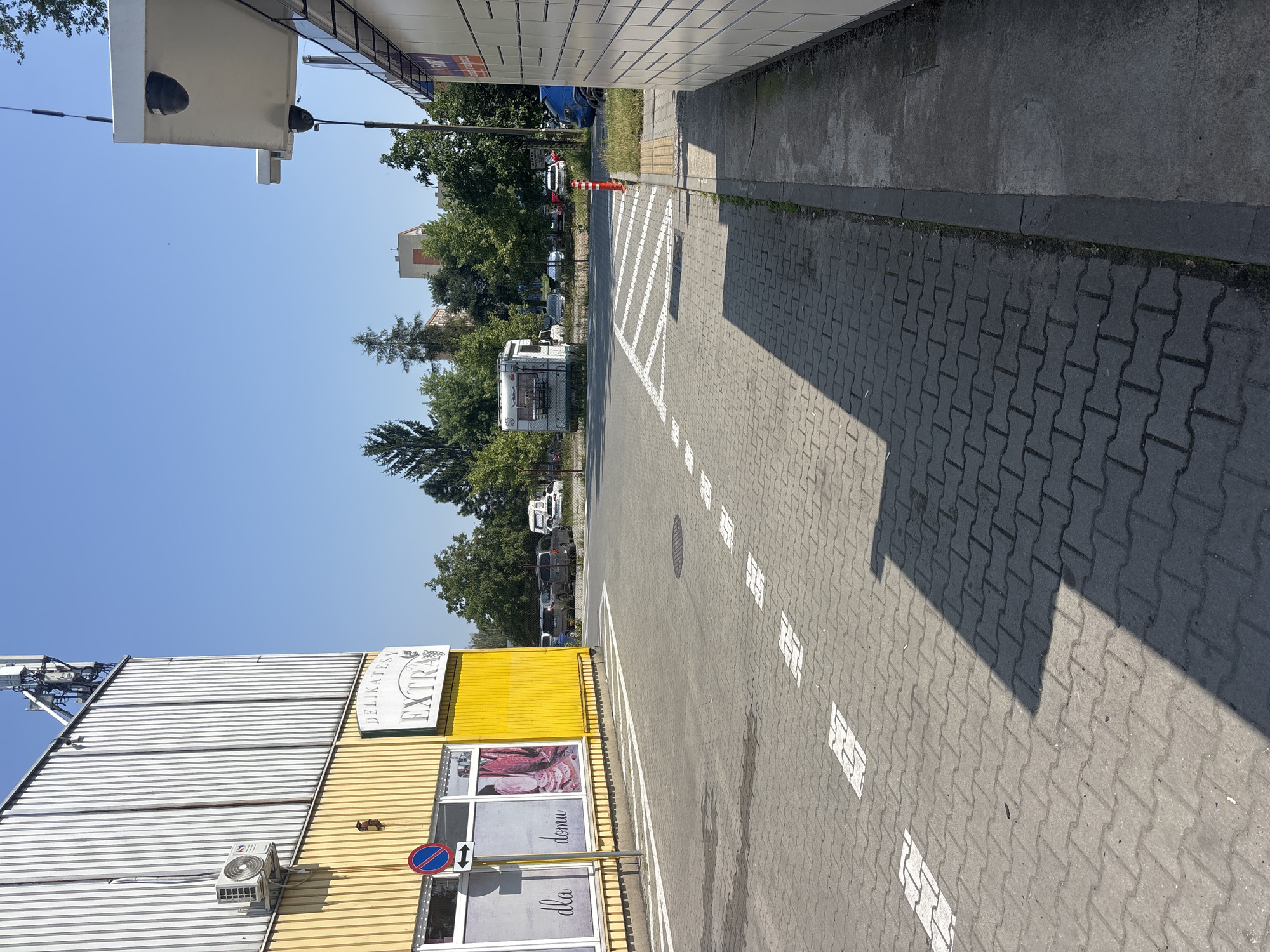 A sunlit, outdoor scene in Krakow, Poland, depicts the exterior of a "DELIKATESY EXTRA" shop. The building features a bright yellow lower section and a corrugated metal upper facade, with an air conditioning unit mounted on the yellow wall. A circular sign with a red border and a blue center, displaying a red diagonal line and a white arrow pointing left, is affixed to the building. A window displays images of meat products and text reading "dla domu."
The ground is paved with interlocking grey bricks, marked with white dashed lines and solid white arrows indicating traffic flow. In the background, several cars are parked in front of trees and buildings. A white RV is also visible. To the right, a modern, white-paneled structure with multiple compartments, possibly a parcel locker system, stands adjacent to the paved area. The sky is clear and blue.