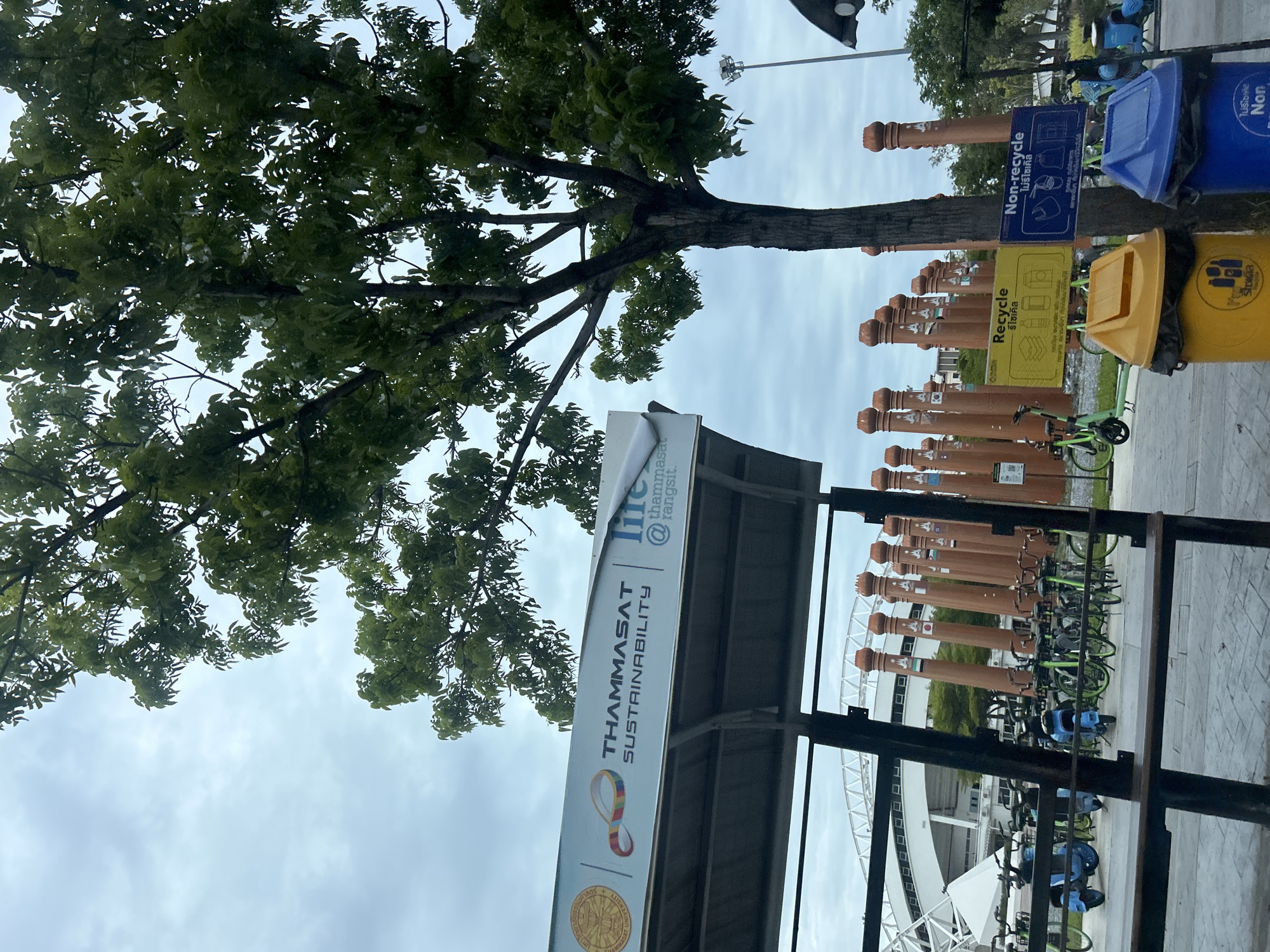 This image captures an outdoor scene, likely within a public area, potentially at a university campus given the prominent "Thammasat Sustainability" signage. In the foreground, a covered seating area with a wooden bench is visible, suggesting a place for rest or waiting. Adjacent to this, a cluster of blue and green electric scooters and bicycles are parked, indicating a mode of transportation.
Dominating the background are rows of decorative, slender orange pillars, each adorned with flags. Beyond these pillars, a modern stadium or large building with white architectural elements is discernible. A large tree with lush green foliage is a significant element, its branches extending across the upper portion of the frame.
Two prominent recycling bins, one yellow and one blue, are situated near the tree, each with corresponding signage in Thai. The yellow bin is for "Recycle" and the blue bin is for "Non-recycle." The sky is overcast with a cloudy, muted sky. The overall impression is one of a well-maintained public space designed for community use, likely in a city in Thailand given the Thai language on the signage.