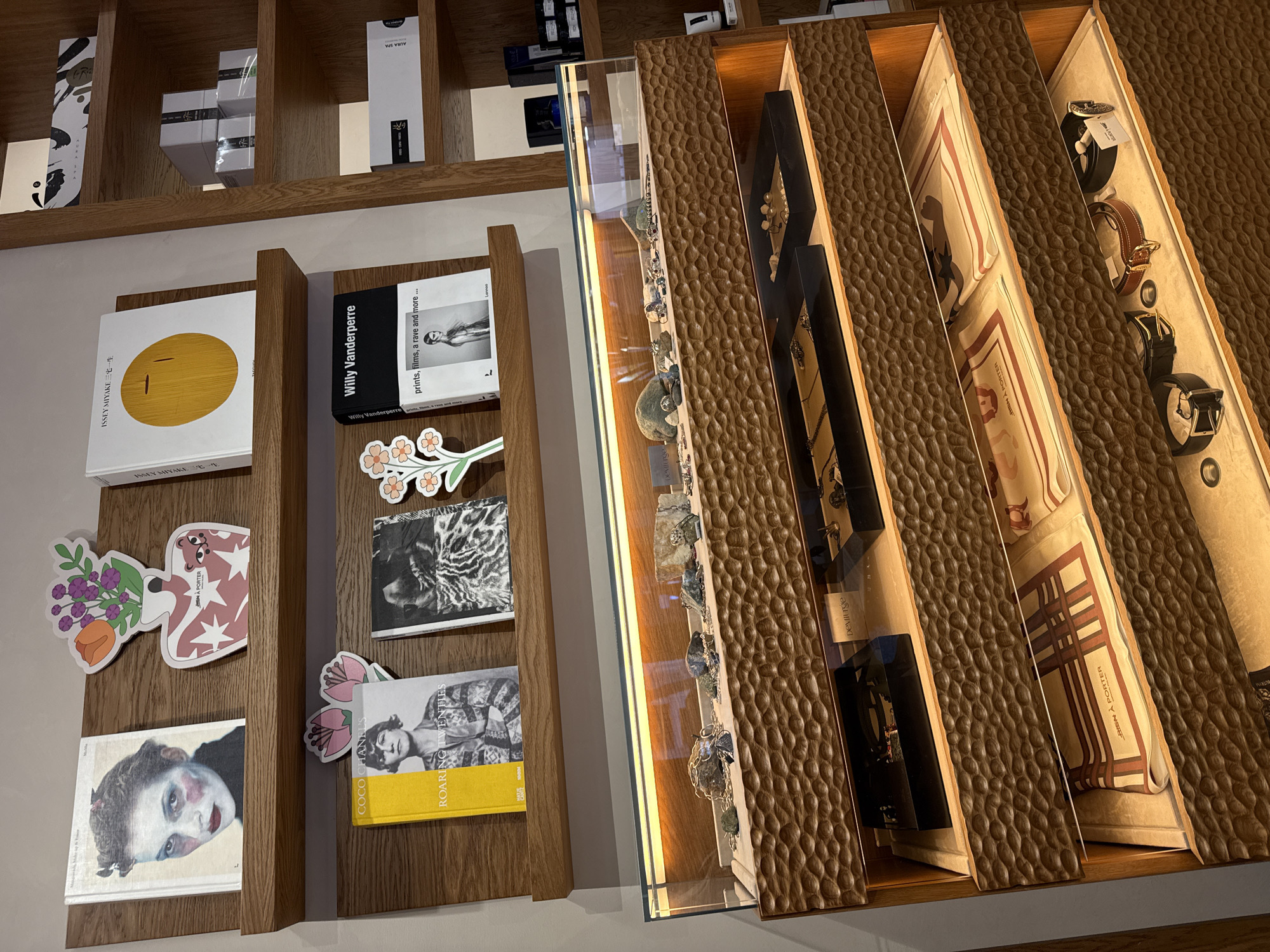This image depicts an interior view of a retail establishment located at 39 Tumanyan Street, Yerevan, Armenia, identified by "BIEN A PORTER" branding on displayed items. The scene is dominated by a combination of wooden shelving units and tiered display cases.
On the upper left, two simple light-colored wooden shelves are mounted against a light grey wall. The top shelf holds three items: a book titled "Making Faces, Makeup & Maniacs" featuring a woman's portrait with red blush, a decorative cutout shaped like a white vase with red stars and a pink bear logo containing purple and orange flowers, and a book titled "ISSEY MIYAKE" with a large yellow circle design. The lower shelf displays three additional items: a yellow-spined book titled "COCO CHANEL'S ROARING TWENTIES" with a portrait of a woman, a second decorative cutout with two pink flowers, and a black-spined book titled "Willy Vanderperre: prints, films, a rave and more..." featuring a black and white photograph of a woman.
To the right, a multi-tiered wooden shelving unit extends vertically, containing various packaged goods. Visible items include stacked grey boxes, white rectangular boxes with text, and smaller dark bottles or containers. One white box is labeled "AURA SPA."
In the lower half of the image, a large, multi-level display unit features a prominent glass-covered top section. This top section contains various natural stones, minerals, and what appear to be necklaces or jewelry laid out on a light-colored surface. A brand label, "DEVIL'TSE," is visible within this case. Below the glass display, the unit consists of three additional tiers of open shelves, each with a dark, textured, carved wooden front resembling repetitive, organic wave patterns. Each tier is illuminated from above by strip lighting. The second tier down from the top glass case holds multiple black rectangular trays containing jewelry, including rings and possibly earrings. The third tier displays folded textiles, identified as "BIEN A PORTER" on the visible fabric. The bottom tier showcases various leather belts and accessories.