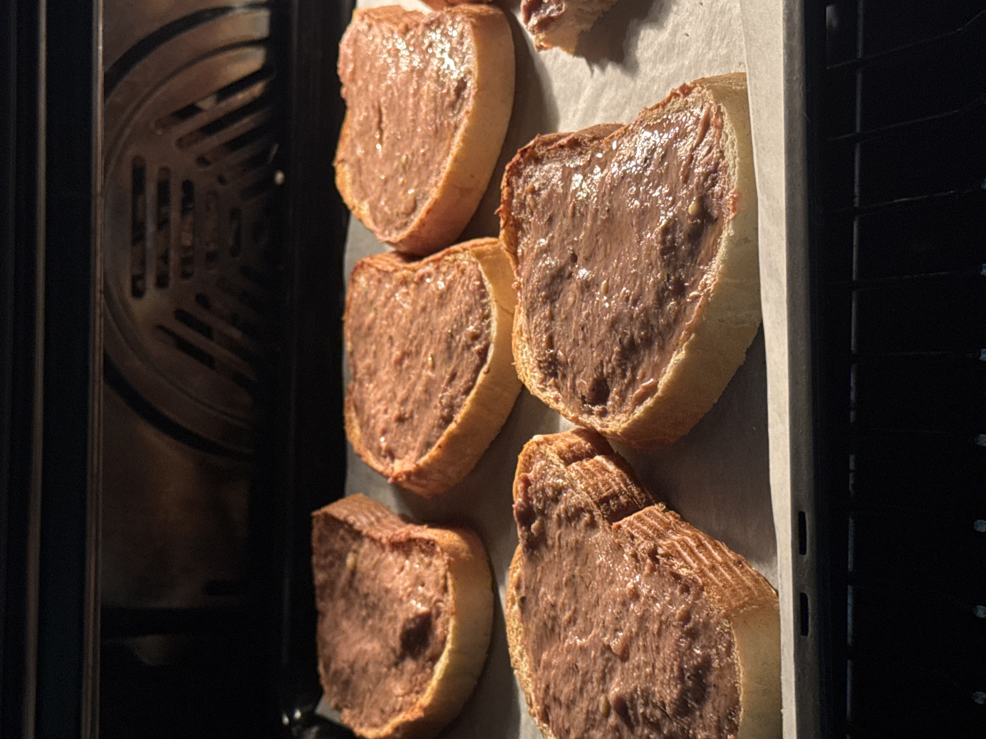 The image shows a baking sheet with five slices of bread spread with a brown paste, likely liver pate, inside an oven. The oven's interior is visible on the left, showing the heating element. The baking sheet is lined with parchment paper. The scene takes place indoors. The location cannot be determined based on the visual elements of the image. There are no people visible in the frame.