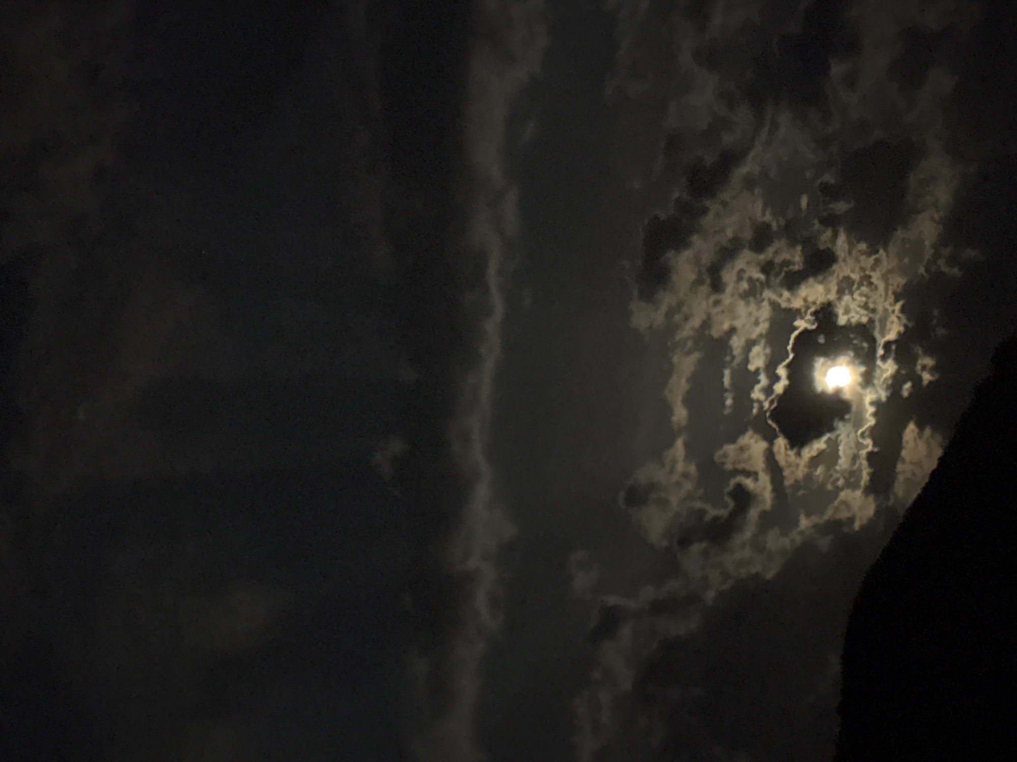 The image displays a nocturnal sky, predominantly dark with varying densities of clouds. A bright, circular light source, identified as the moon, is positioned towards the upper right side of the frame. It is partially veiled by an intricate arrangement of clouds, which are illuminated from behind, creating glowing edges and swirling patterns where the moonlight penetrates. These illuminated cloud formations stand out against the darker, unlit areas of the sky.
A prominent, dark, silhouetted mass occupies the far right edge of the image, extending vertically. This feature suggests a natural landform such as a mountain or hill, contributing to the dramatic composition of the scene. The clouds on the left and upper portions of the image appear denser and darker, gradually becoming more translucent and brightly lit closer to the moon.
The scene captures a natural landscape at night, characterized by the interplay of moonlight and clouds. No human activity, individuals, or man-made objects are visible. The weather appears cloudy. There is no visible text. The provided context indicates the location as Budva, Montenegro.