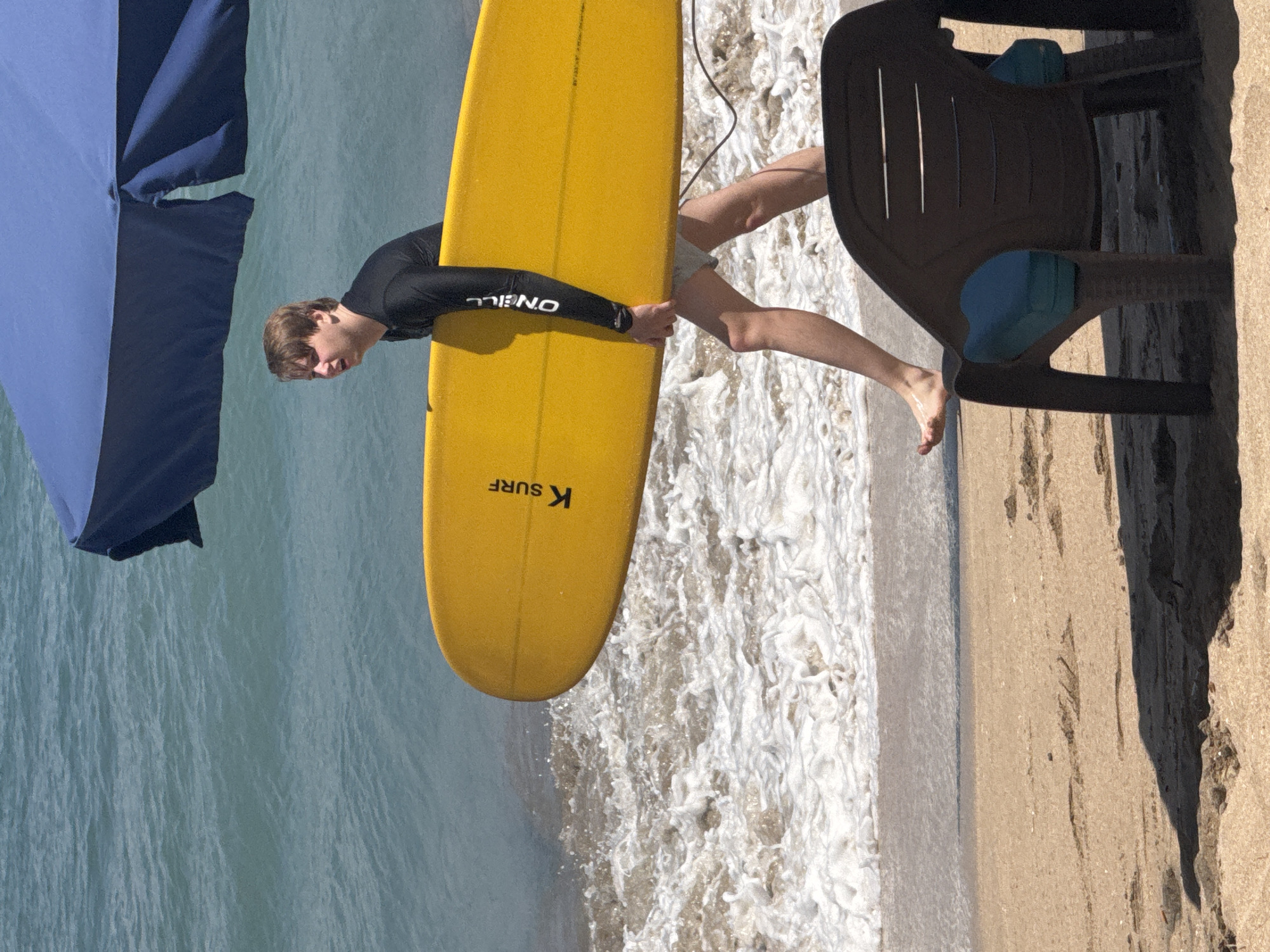 A young Caucasian male wearing a black long-sleeved rash guard and light-colored swim shorts walks from the ocean towards the beach carrying a yellow surfboard under his right arm. The surfboard has "K SURF" and "O'NEILL" branding visible. He is partially shaded by a blue umbrella. The ocean waves are breaking on the sandy shore. In the foreground, a dark brown plastic beach chair with blue cushions sits on the sand, casting a shadow. The scene is a beach in Kuta, Indonesia.