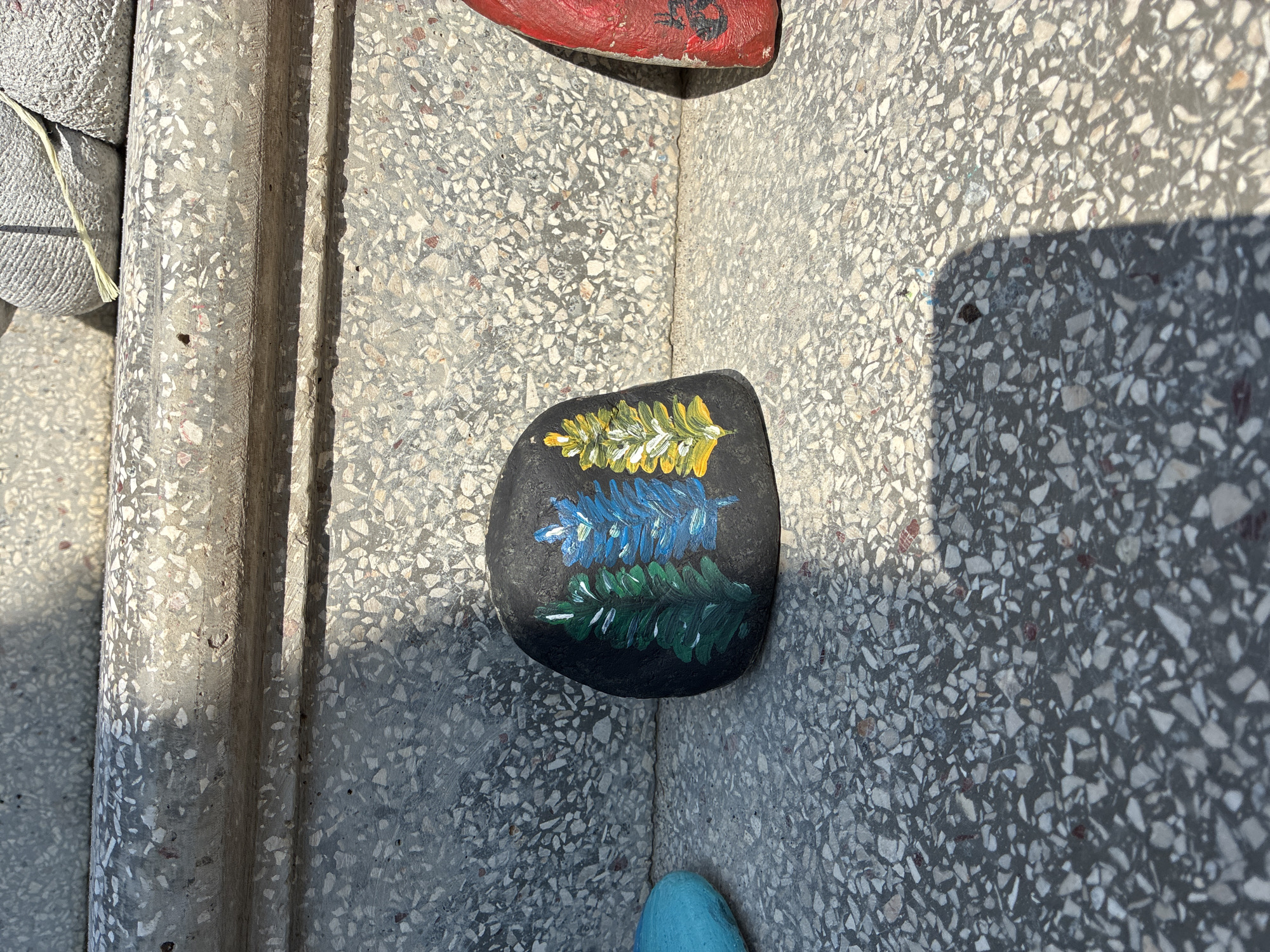 A close-up, eye-level shot shows a painted rock with three stylized evergreen trees on a terrazzo-patterned surface, likely steps or a pathway, in Surami, Georgia. The rock is dark gray and centrally placed, with a green tree on the left, a blue tree in the middle, and a yellow and white tree on the right. A bright turquoise rock is partially visible to the left, and a red rock with a dark design is to the right. The background consists of concrete steps with a terrazzo aggregate of light gray, white, and reddish-brown chips. Shadows from an unseen object fall across the scene, indicating it is outdoors during daylight.