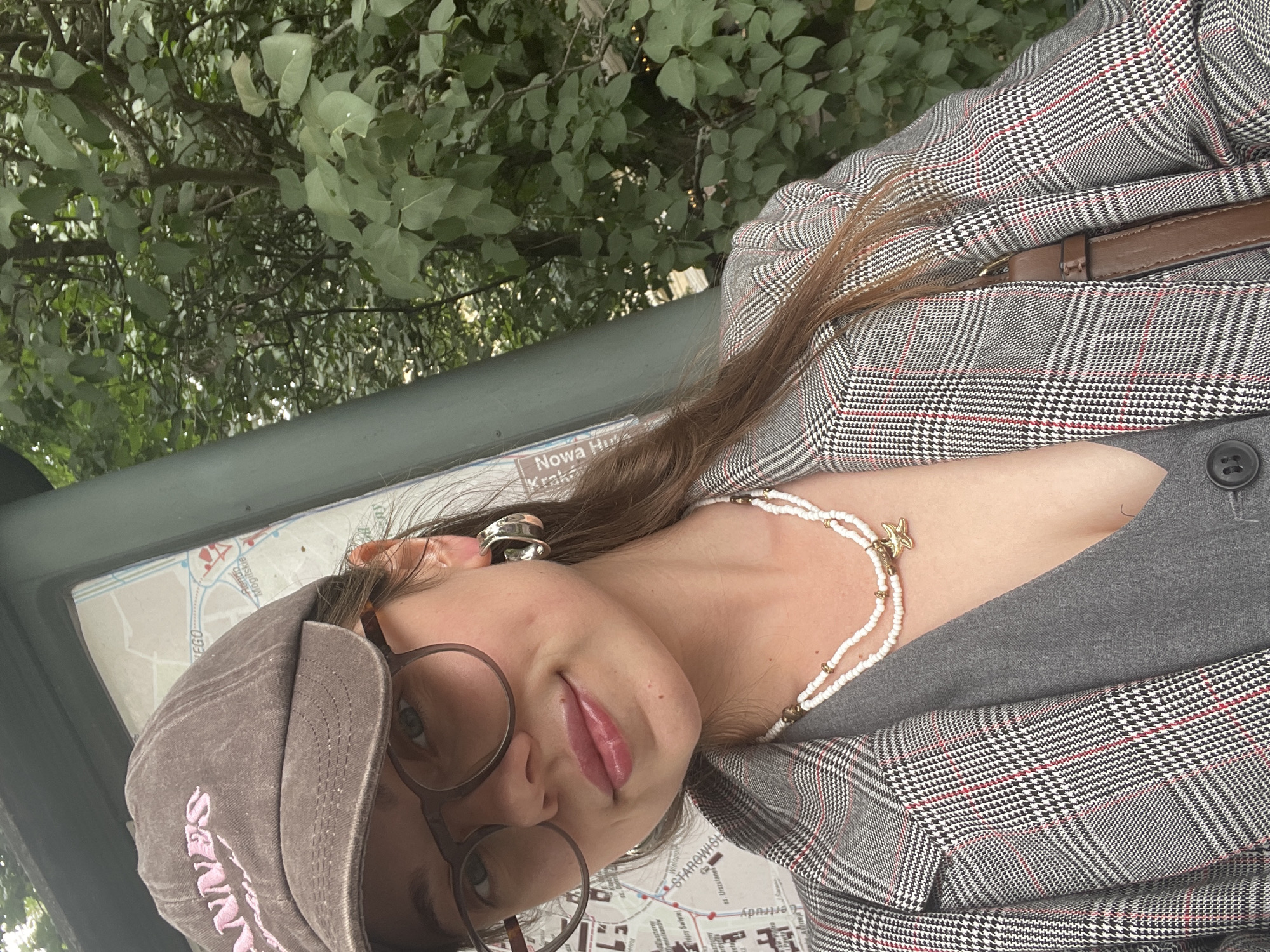A young woman is taking a selfie outdoors. She is wearing a brown baseball cap with pink lettering, glasses, large silver hoop earrings, a grey vest, and a grey and black plaid blazer. She has a two-strand beaded necklace with a starfish charm. Behind her is a map on a green post and lush green trees. The map appears to be in Krakow, Poland, as it contains Polish text such as "Starowislna" and "Nowa Huta".