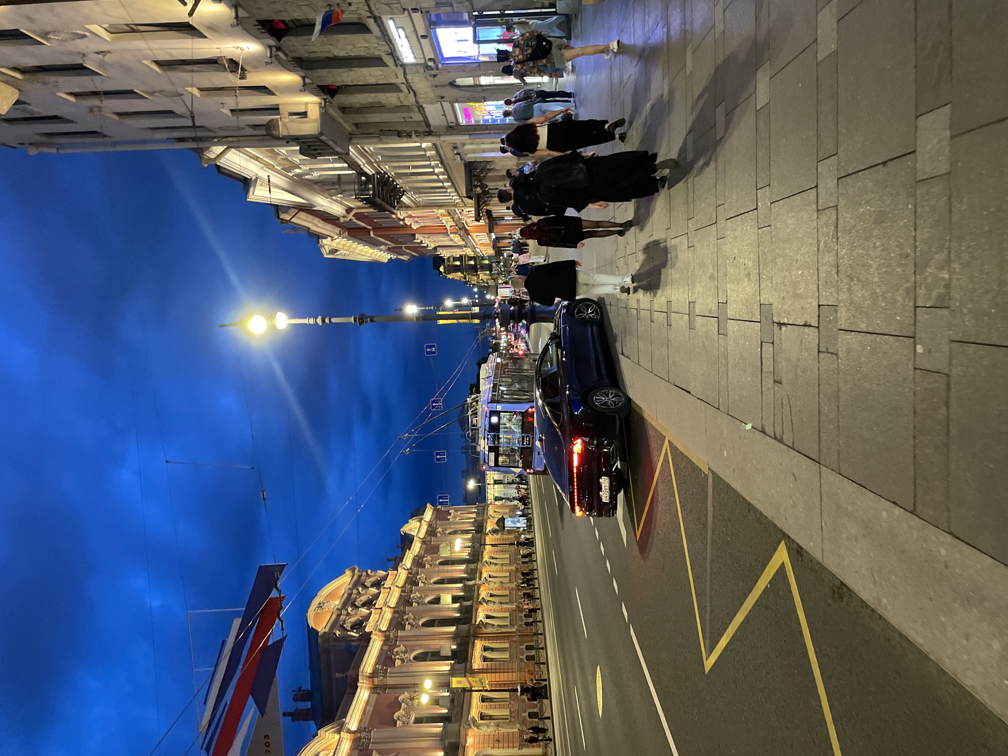 This image depicts an urban street scene at twilight or night on Nevsky Prospekt in Saint Petersburg, Russia. The sky is a deep, dark blue, indicating the evening hours.
On the left, a large, ornate Baroque-style building, identifiable as the Stroganov Palace, dominates the view. Its pale orange/pink facade, characterized by numerous arched windows, columns, and decorative stucco work, is warmly illuminated by lights from below. A decorative banner, striped in red, white, and blue, with the partial number "703" visible, hangs across the street above this building.
The wide asphalt street features white dashed lane markings. On the right side, a yellow zig-zag line indicates a no-parking zone. A dark blue sedan, a Kia K5 (identifiable by its taillights), with a Russian license plate "0475XT 198 RUS" (indicating the Saint Petersburg region), is parked facing away from the camera on the right curb, its rear lights illuminated. Further down the street, a blue and white trolleybus is partially visible in motion, moving away from the viewer under a network of overhead electrical wires.
On the right side of the street, a row of multi-story historical buildings houses various commercial establishments on their ground floors, which are brightly lit with display windows. Several small Russian flags (horizontal white, blue, and red stripes) are affixed to these buildings. An ornate, tall streetlight with a bright yellow glow stands in the mid-ground.
The sidewalk on the right, paved with rectangular stone slabs, is occupied by a group of approximately ten pedestrians walking away from the camera. Their attire is varied, including dark coats, casual tops, shorts, and skirts. Additional pedestrians are visible in the distance on both sidewalks.
