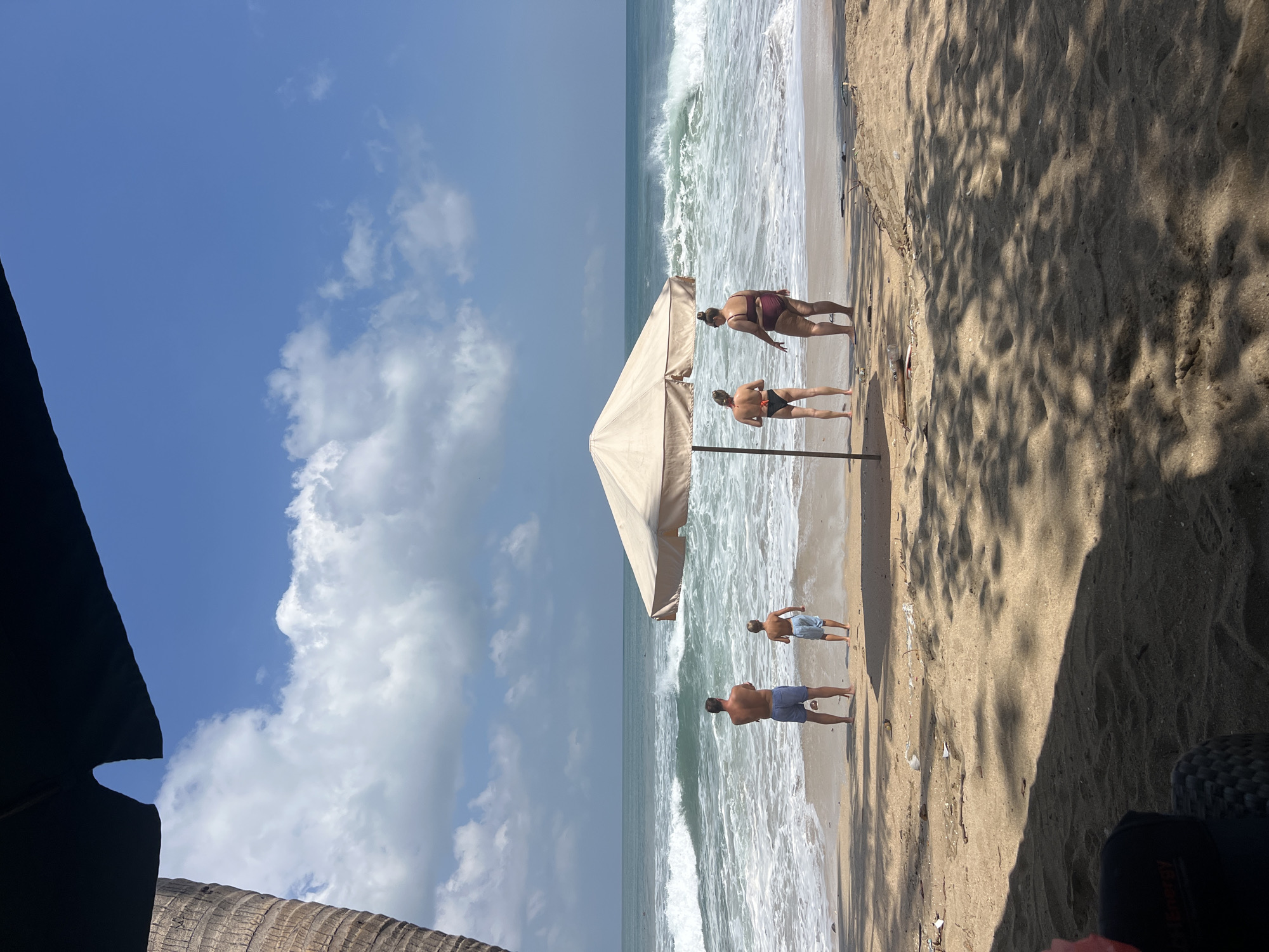 A group of four people are standing on a sandy beach in Kuta, Indonesia, facing the ocean. Two adults, a man and a woman, and two children, a boy and a girl, are positioned near the shoreline, under a large, tan-colored umbrella. The man and boy are closer to the water, while the two women are situated behind them, closer to the umbrella. The waves are crashing on the shore, indicating a strong current or high tide. The sky is clear blue with scattered white clouds, and a palm tree trunk is visible on the left side of the frame. The sand is littered with small pieces of debris, and shadows of leaves are cast across the ground from an unseen tree. In the foreground, a dark-colored object, possibly a bag or chair, is partially visible. The scene appears to be a typical beach day with people enjoying the ocean view.