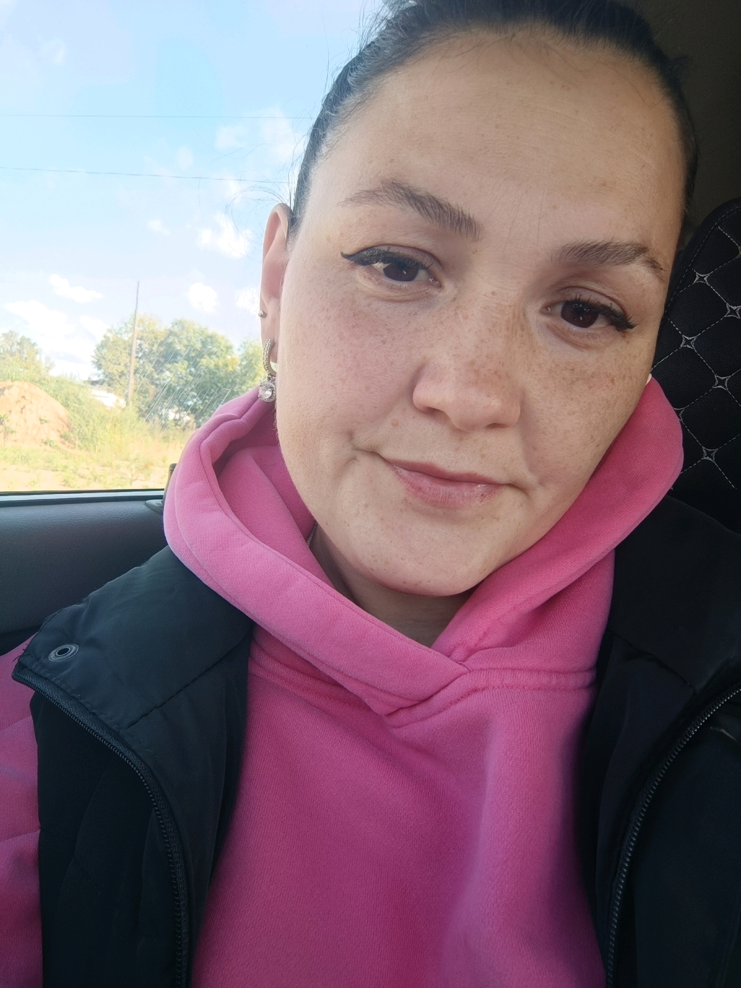 The image is a close-up, selfie-style shot of a woman with fair skin, freckles, and dark hair pulled back. She is wearing a pink hoodie and a black vest or jacket. She has on eye makeup and a large, sparkly earring. The background shows a blue sky with some clouds, trees, and a dirt mound, suggesting an outdoor or roadside location. The woman is inside a vehicle, as indicated by the car door and seat visible in the frame. The image was taken on a sunny day.